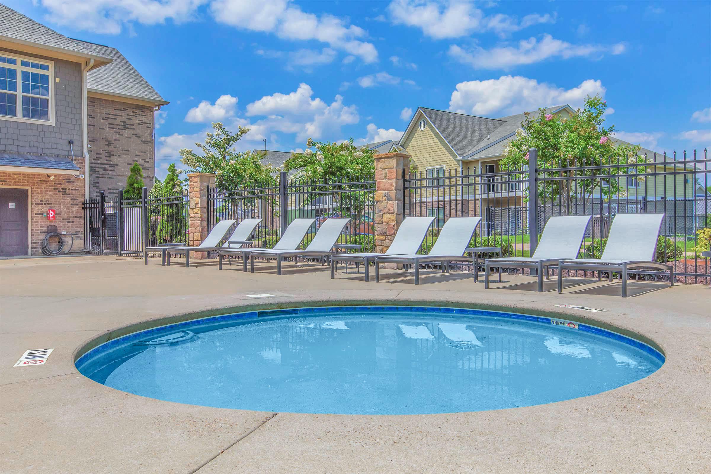 A round hot tub beside a swimming pool area with several lounge chairs arranged neatly around it. The background features residential buildings with fences and greenery under a bright blue sky with scattered clouds.