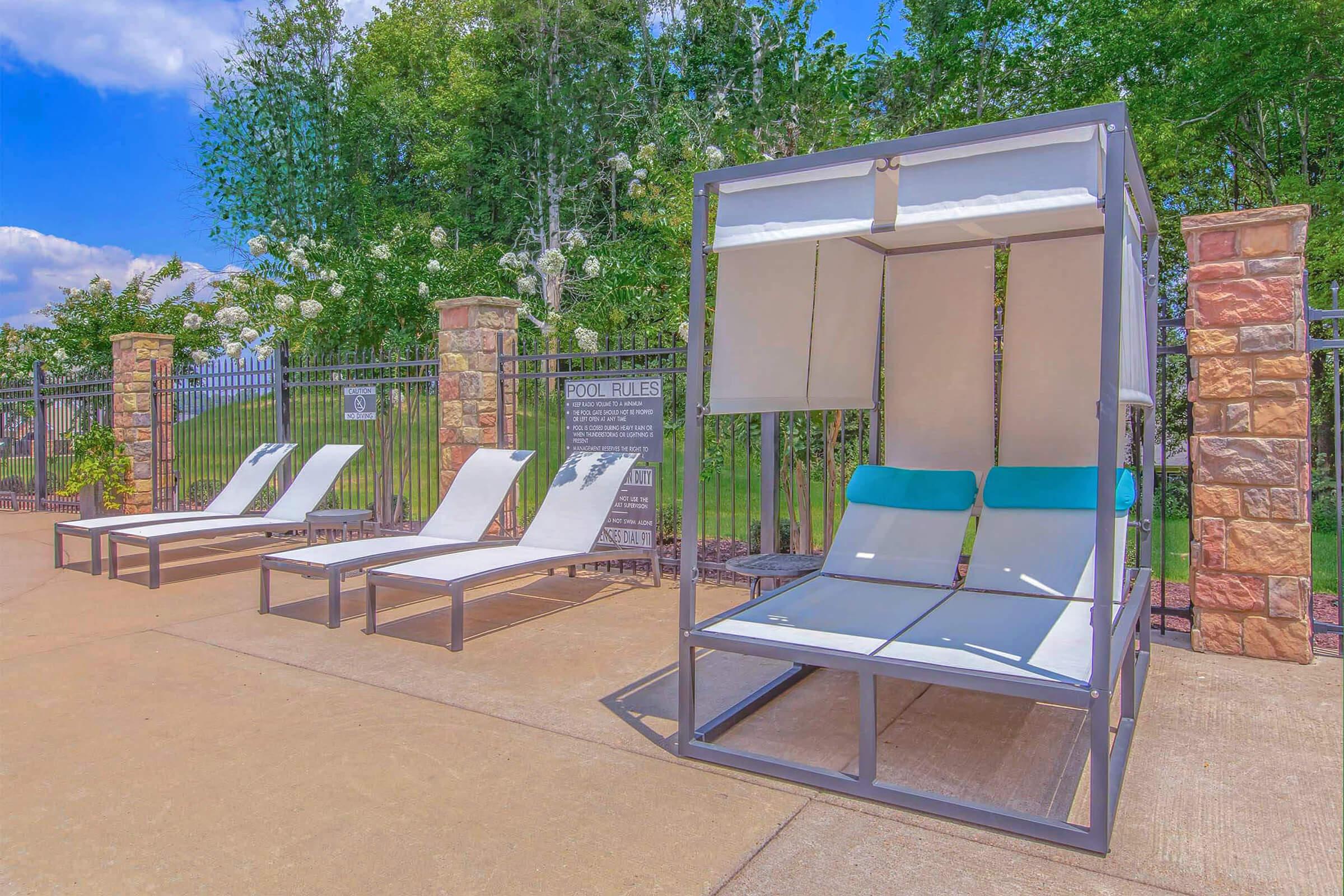 Two modern lounge chairs and a shaded canopy bed are positioned beside a swimming pool area. Lush greenery and flowering shrubs are visible in the background, under a clear blue sky.