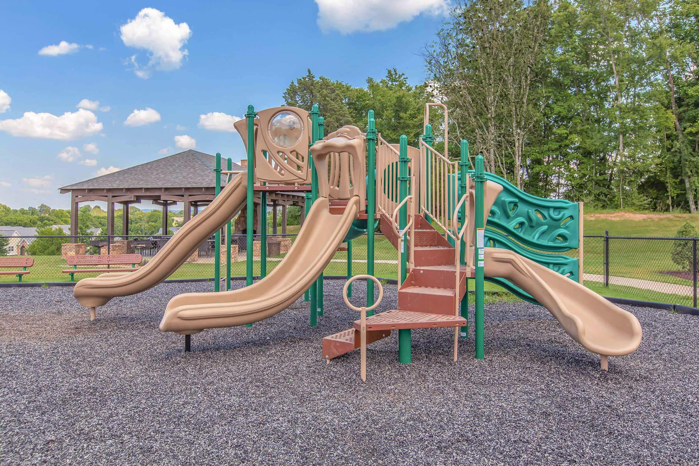 Playground equipment featuring two large slides, a climbing structure, and a small tower, set on a gravel surface with a grassy area and a picnic shelter in the background.