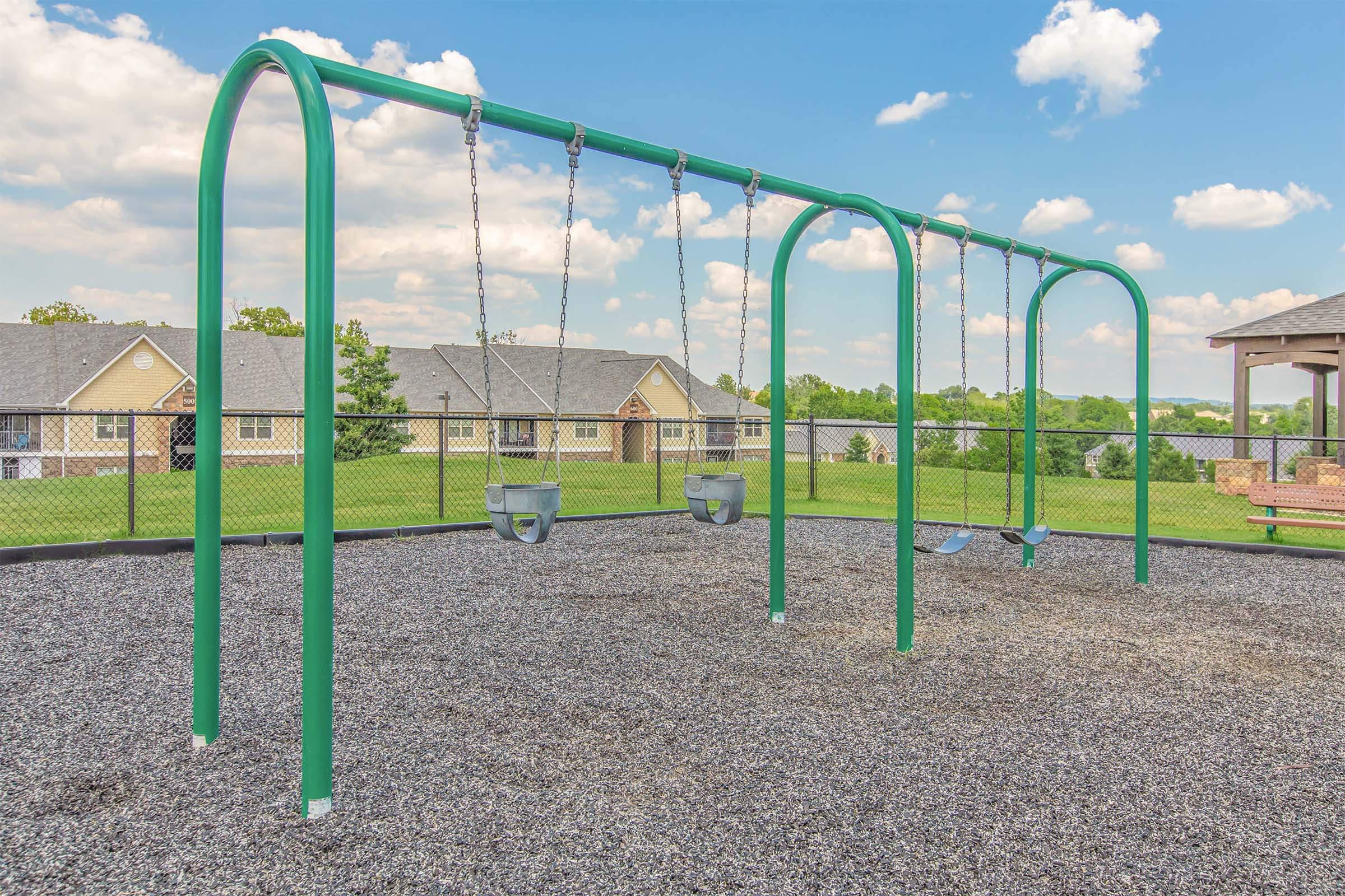 A playground featuring a set of swings with green frames, positioned on a gravel surface. In the background, there are houses and a clear blue sky with fluffy clouds.