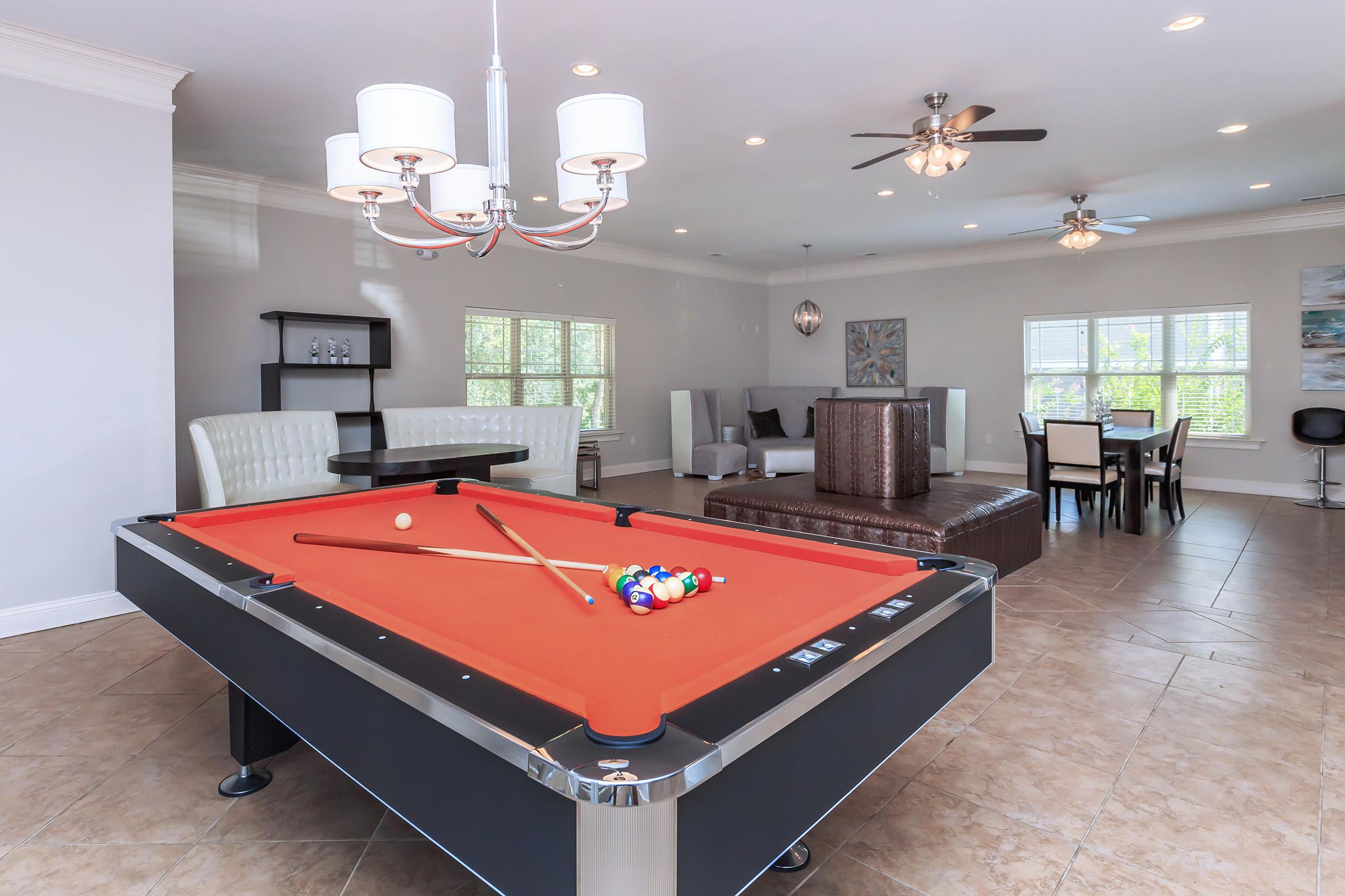 A spacious living area featuring a pool table with an orange felt top in the foreground, a stylish brown leather couch, a dining table, and modern light fixtures overhead. Large windows allow natural light to fill the room.