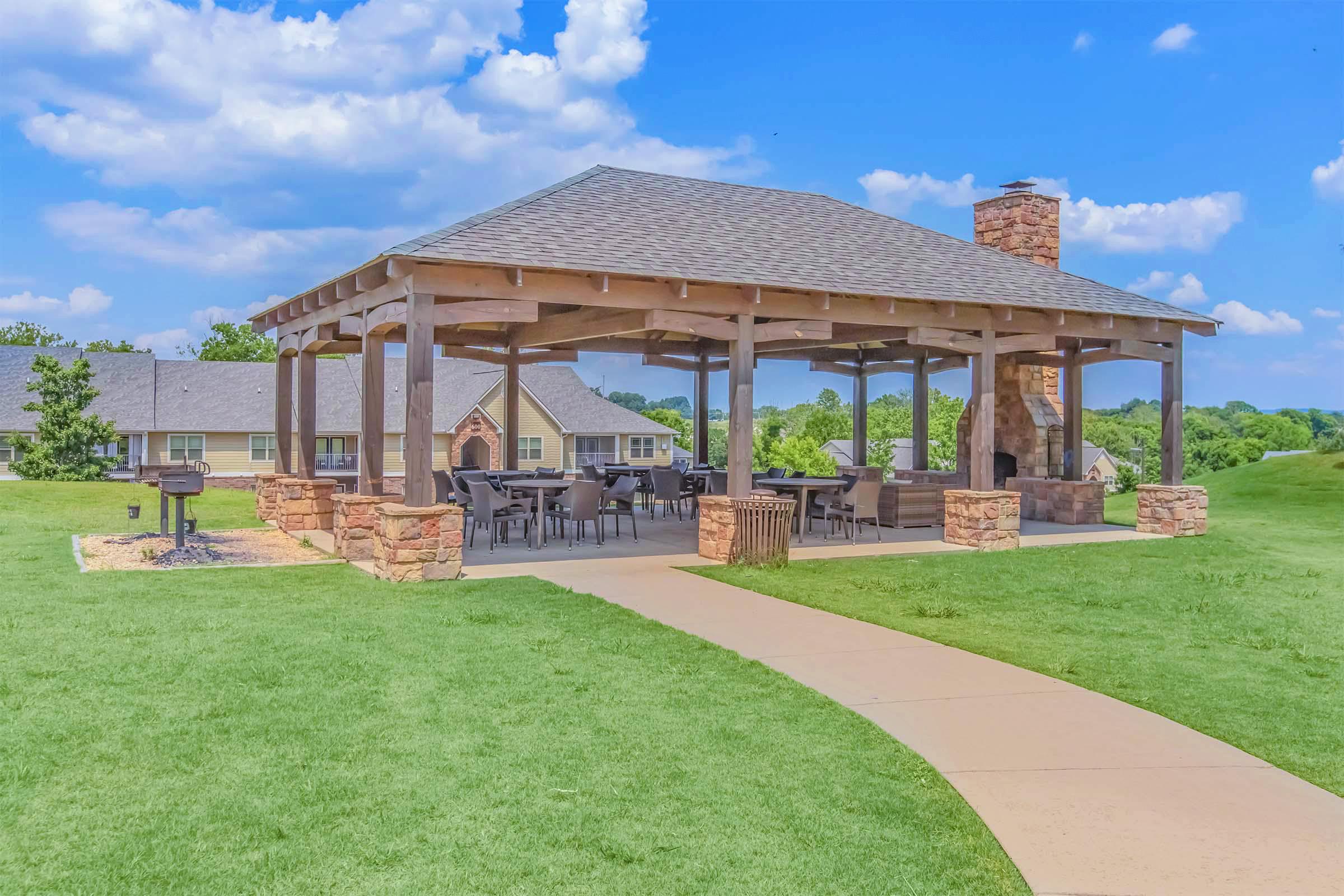 A covered outdoor pavilion with a stone chimney, surrounded by green grass and a walking path. There are tables and chairs underneath the pavilion, and a barbecue grill nearby. The sky is blue with fluffy white clouds.