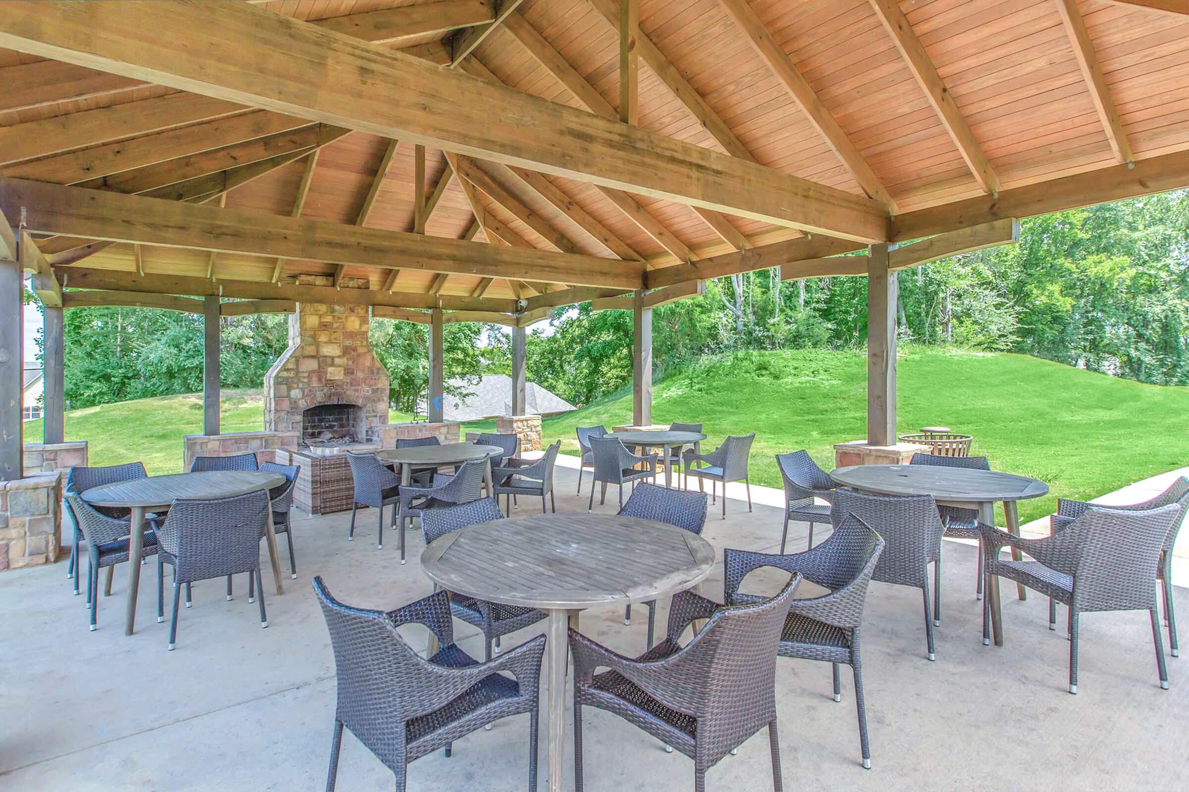 Outdoor pavilion with a wooden roof supported by beams, featuring several round tables and wicker chairs. A stone fireplace is visible in the background, surrounded by green grass and trees.