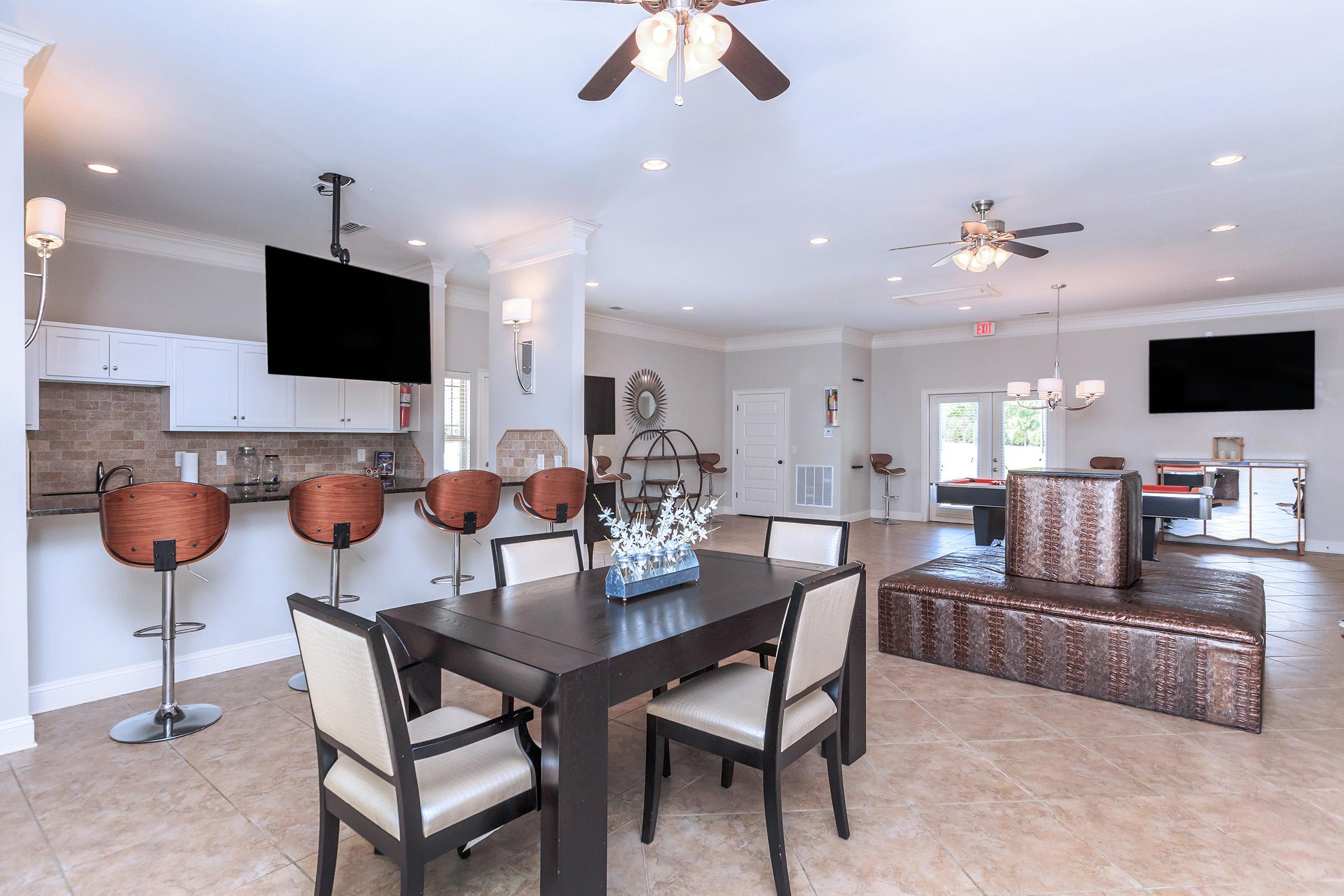 A modern, spacious common area featuring a dining table with four chairs, a sleek bar with stools, and a large sofa. The room has light-colored walls, ceiling fans, and multiple large windows providing natural light. Two flat-screen TVs are mounted on the walls.