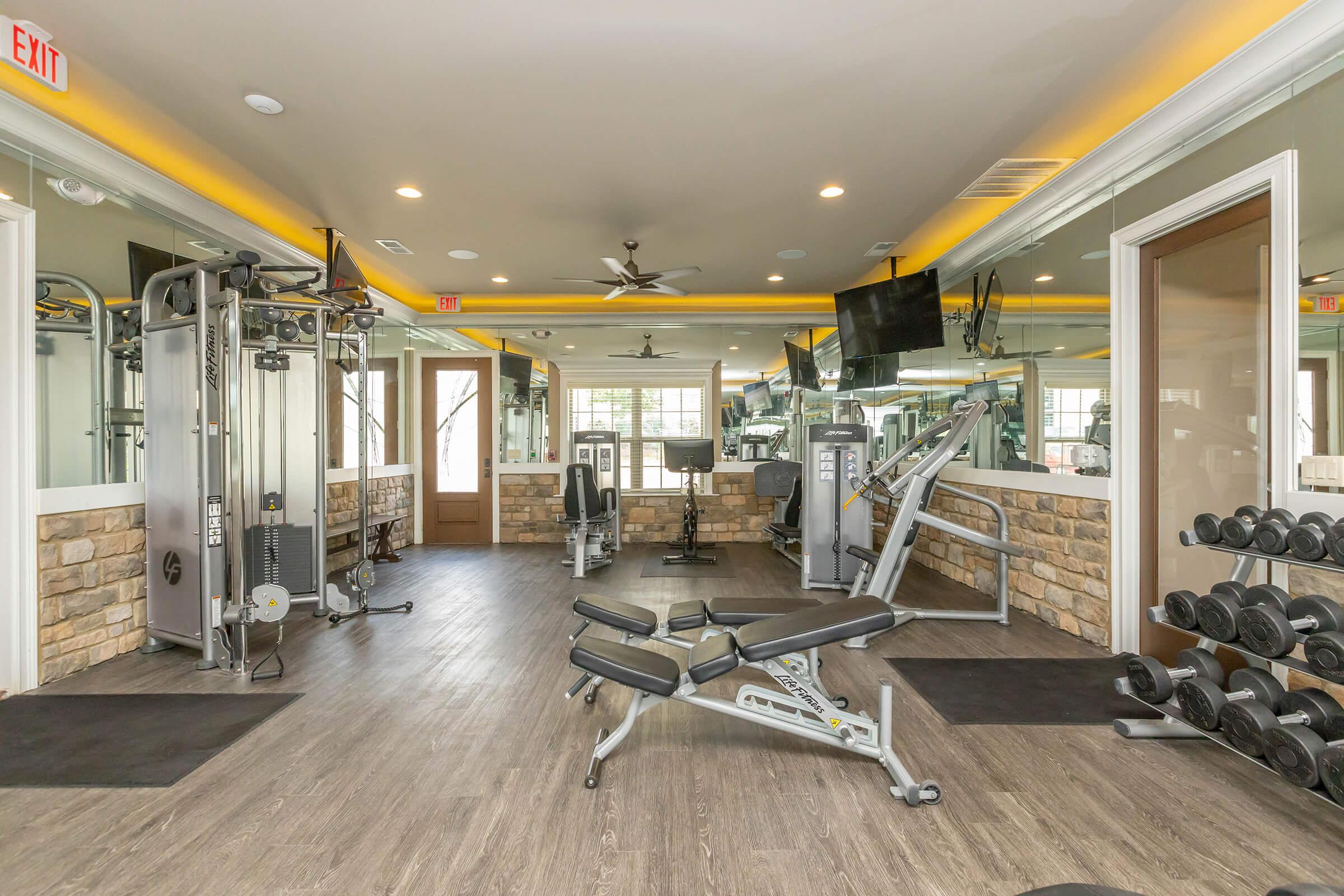 A modern gym interior showcasing exercise equipment such as weight machines, free weights, and a workout bench, with large windows allowing natural light to fill the space. The walls are decorated with stone accents, and there are multiple mirrors and televisions mounted on the walls.