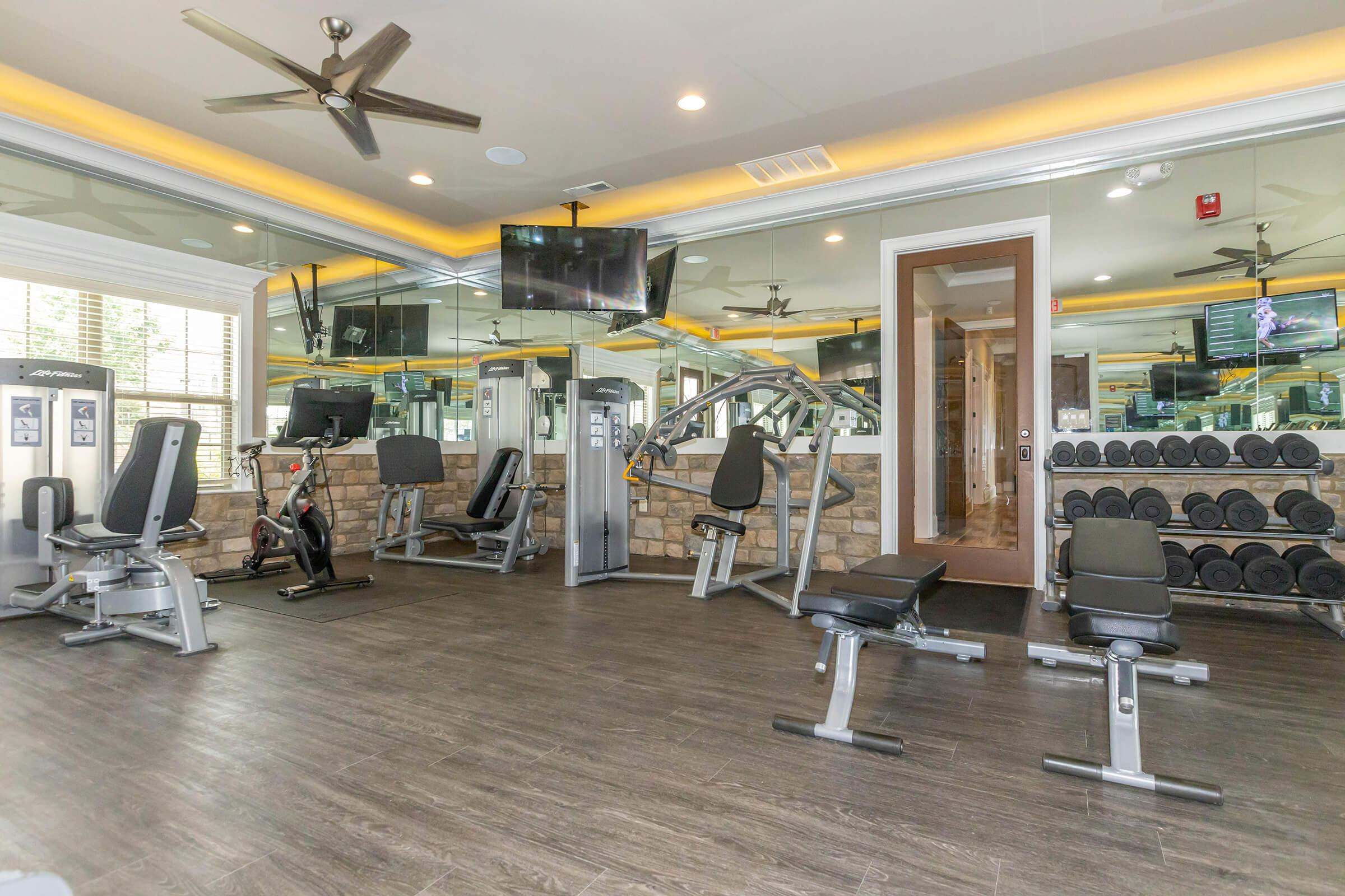 A modern gym interior featuring various exercise equipment, including weight machines, benches, and dumbbells. The space is well-lit with large mirrors and ceiling fans, creating an inviting workout environment.