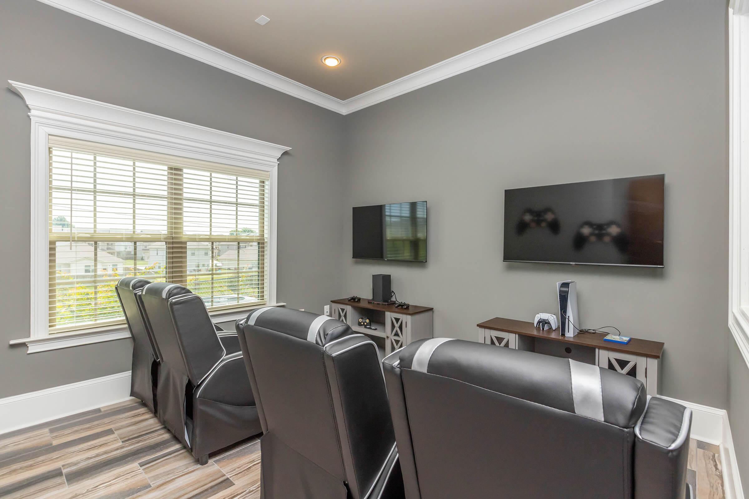 A cozy game room featuring two large wall-mounted televisions, a modern gaming console, and four black reclining chairs arranged in front of the screens. Natural light streams in through a window, and the walls are painted a soft gray.