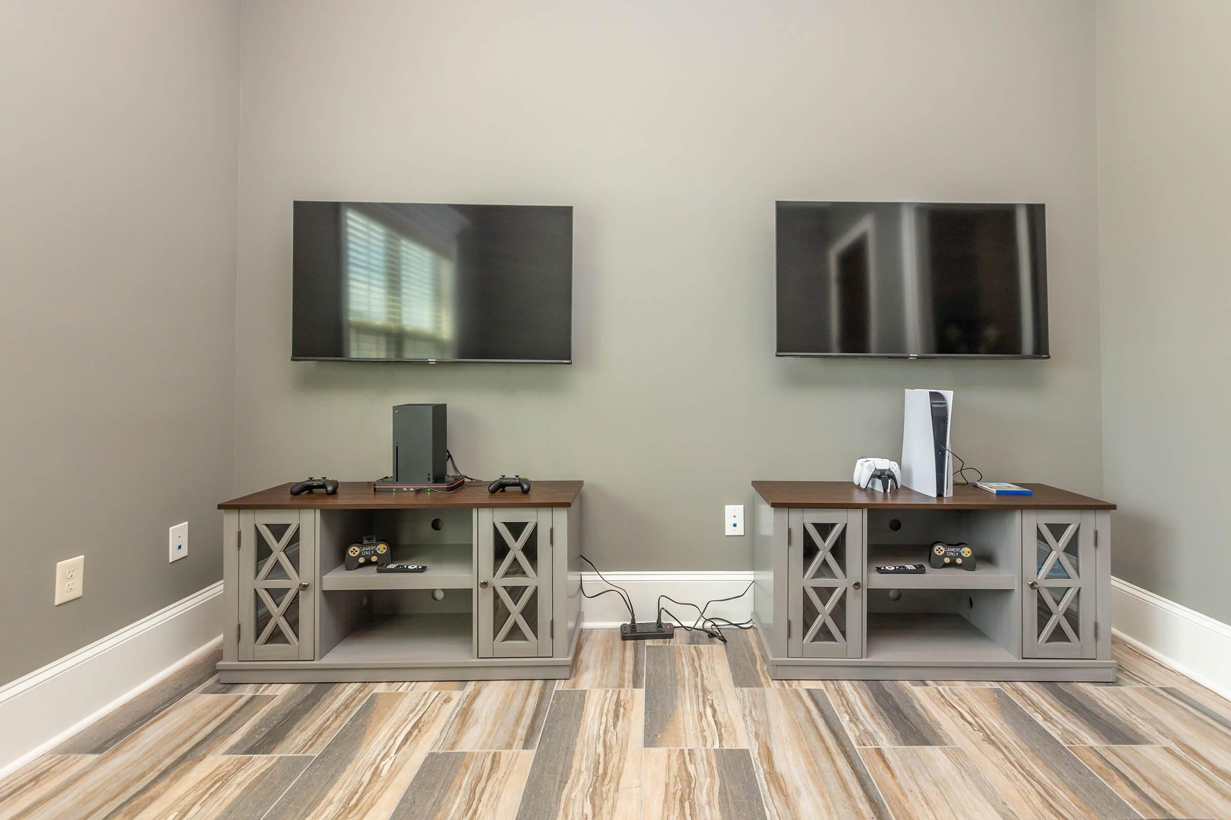 Two entertainment centers with flat-screen TVs mounted on the wall, each featuring a gaming console (one appears to be a PlayStation 5 and the other a different console), game controllers, and other accessories. The walls are painted light gray, and the flooring has a wooden appearance.