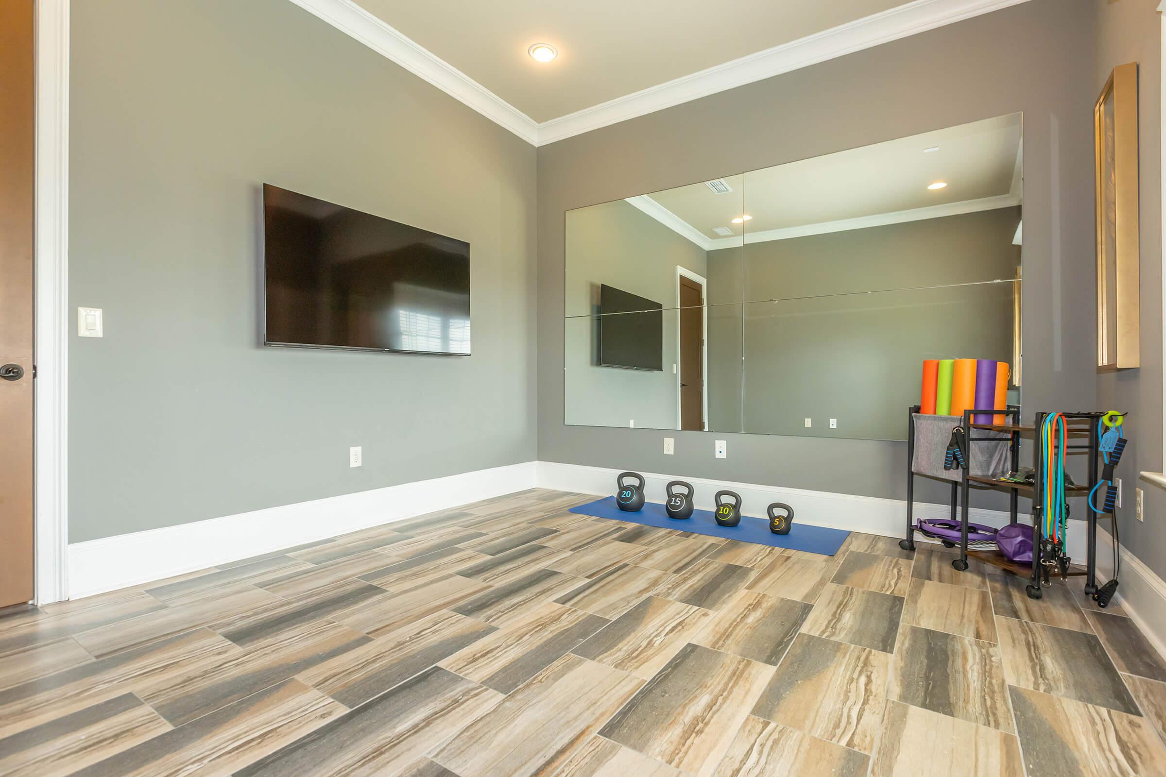 A modern home gym with gray walls, featuring a wall-mounted TV, a large mirror, and exercise equipment including dumbbells and resistance bands on a mat. The flooring is a light wood look tile.