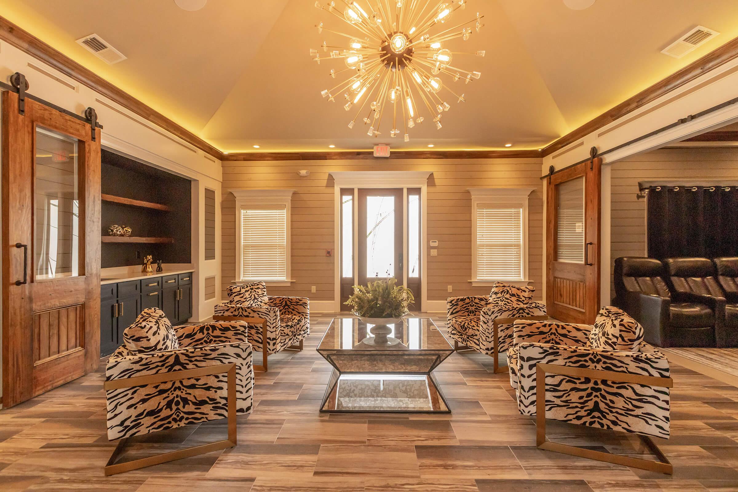 A stylish interior room featuring modern decor with four zebra-print chairs arranged around a glass coffee table. The space has wooden paneling, a large chandelier with multiple light bulbs, and large windows allowing natural light. There is a doorway leading outside, and a cozy atmosphere is created by the warm lighting.