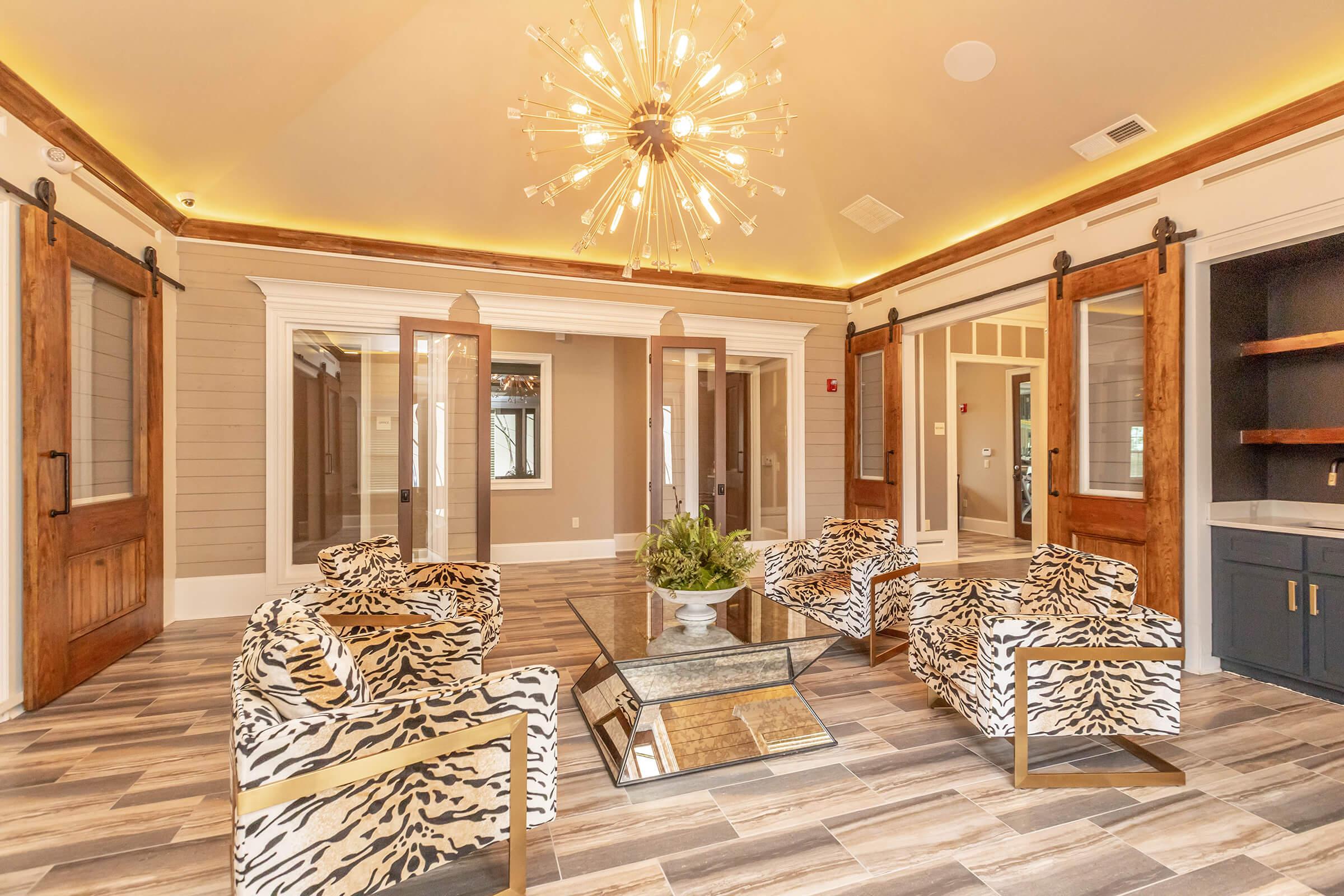 A stylish lounge area featuring modern decor with a large chandelier, zebra-print chairs, a glass coffee table, and wooden sliding doors. The walls are painted in neutral tones, and the flooring is a combination of tiles that resemble wood.