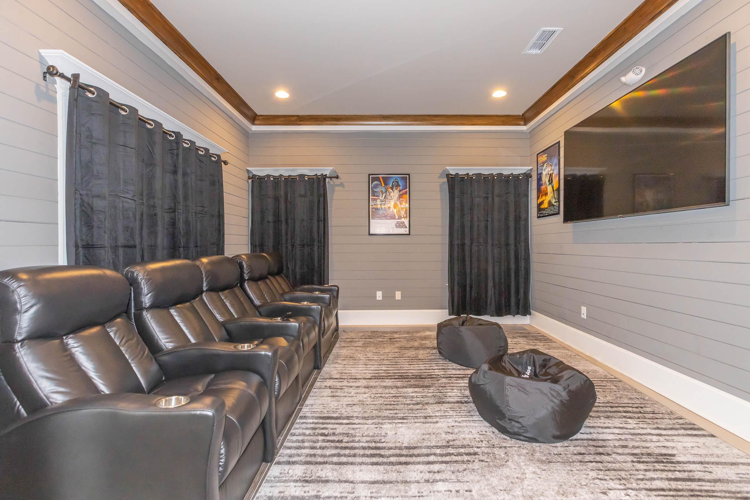 Home theater with black reclining chairs, dark curtains, a large wall-mounted TV, and framed movie posters on the walls. The room features gray wood paneling and a plush area rug.