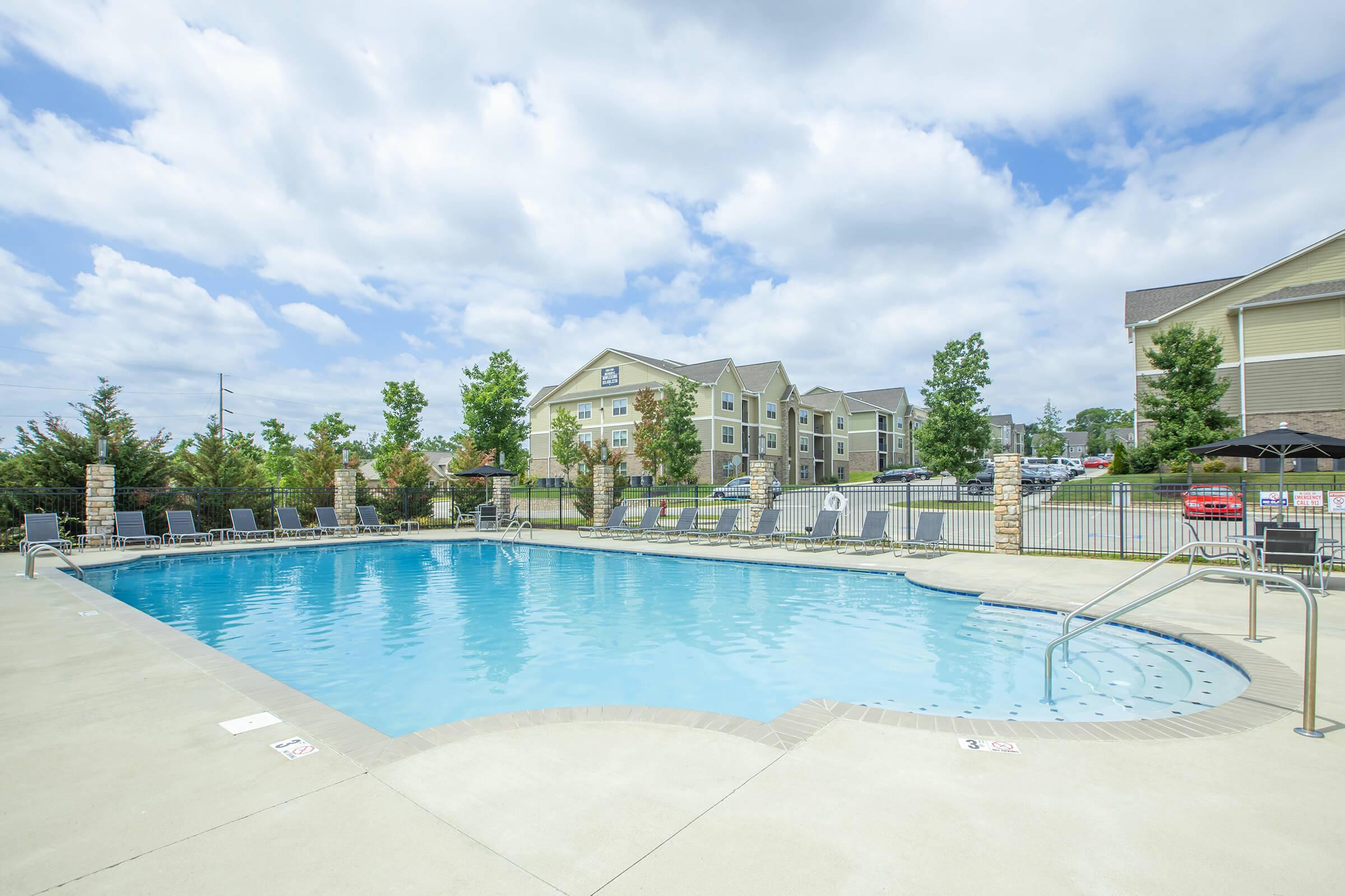 A swimming pool surrounded by lounge chairs, with trees and a multi-story apartment building in the background under a partly cloudy sky.