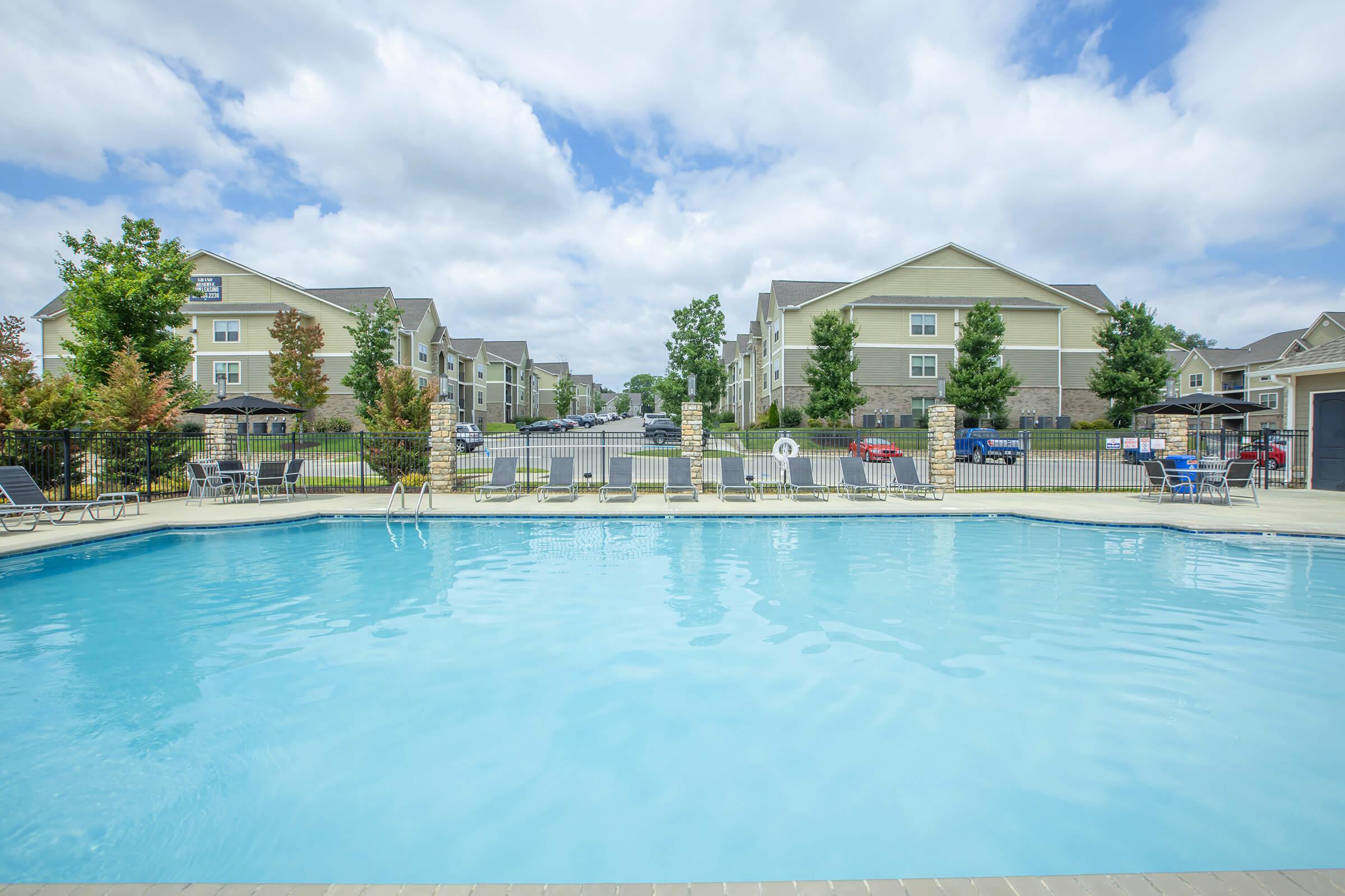 A clear blue swimming pool surrounded by lounge chairs, accompanied by lush greenery and multi-story residential buildings in the background under a cloudy sky.