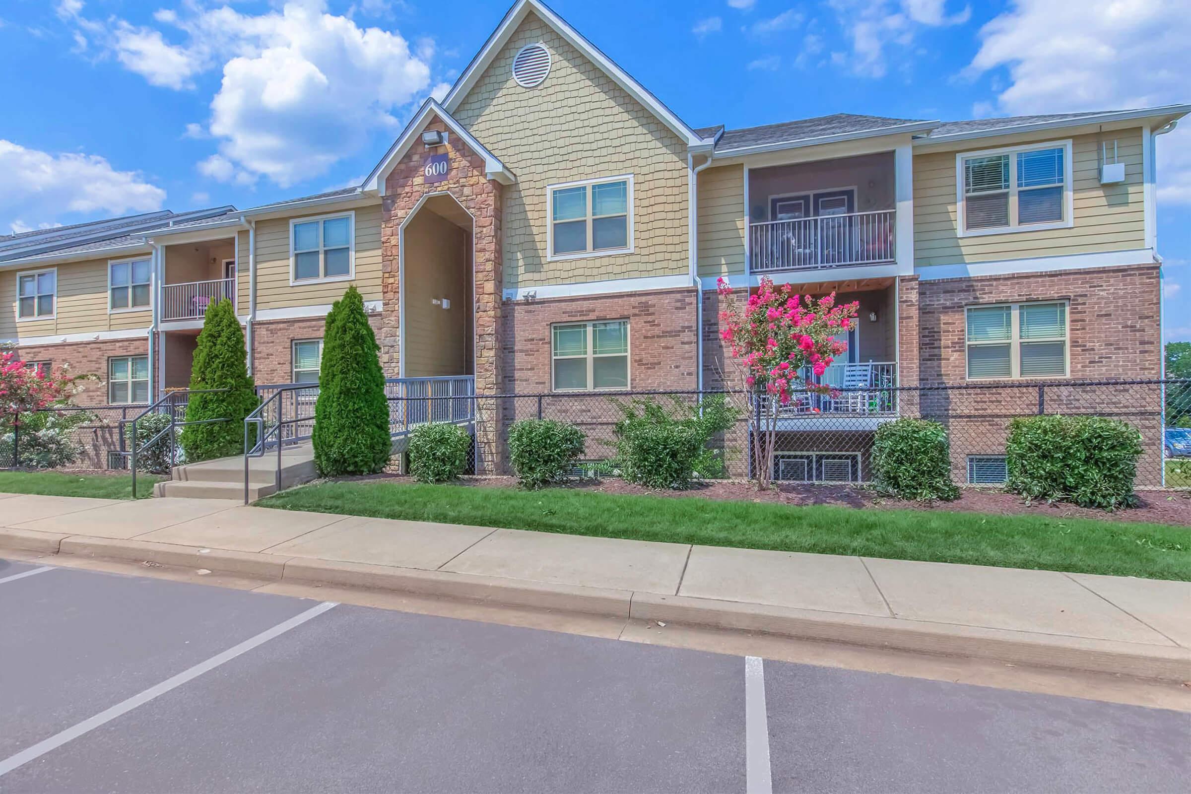 A two-story apartment building with yellow and brick siding, featuring a central entrance and balconies above. Lush green landscaping with small trees and shrubs is present in front of the building, and there are clear blue skies with a few clouds overhead.