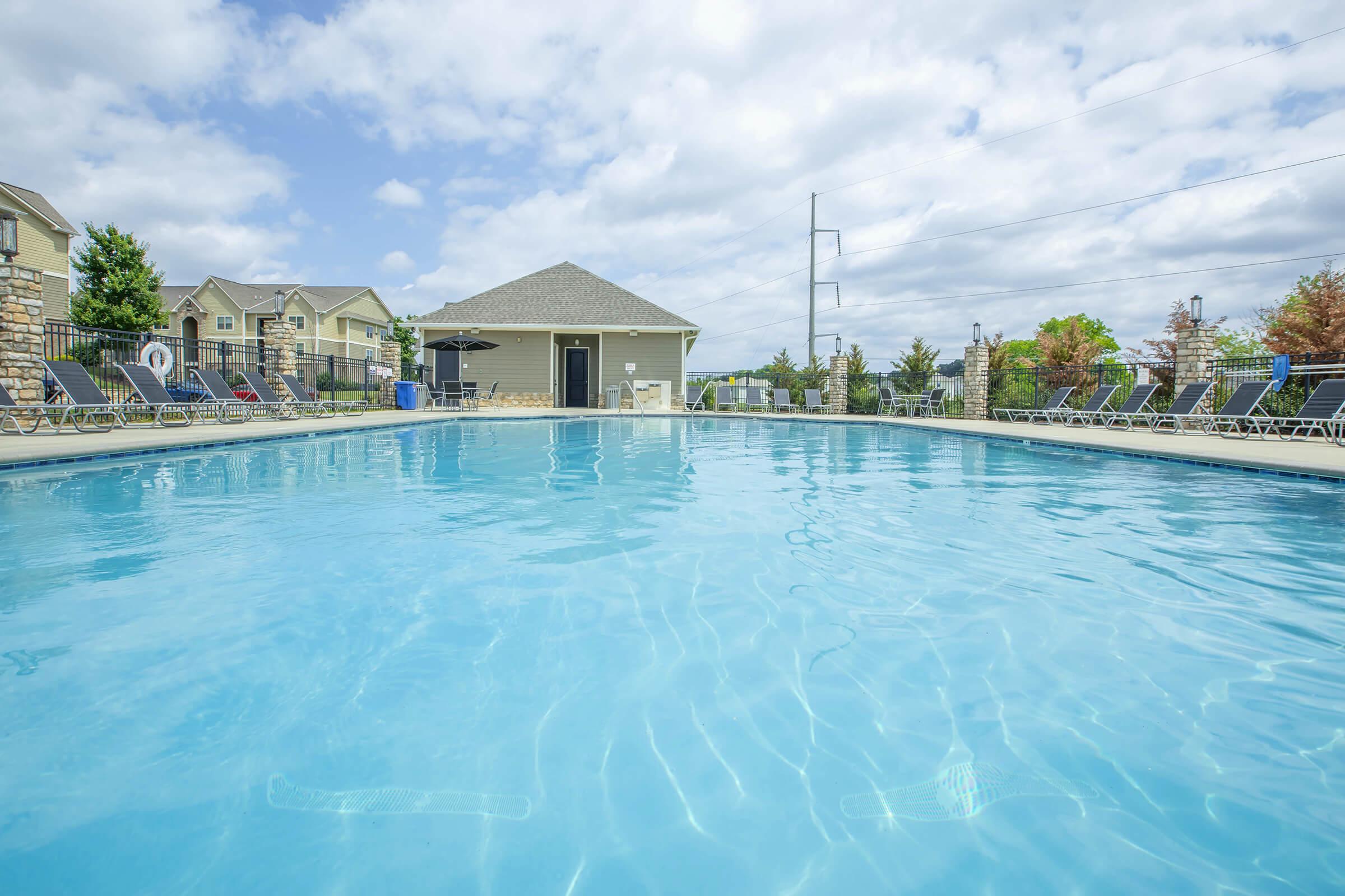 A clear blue swimming pool surrounded by lounge chairs, with a clubhouse in the background and partially cloudy skies.