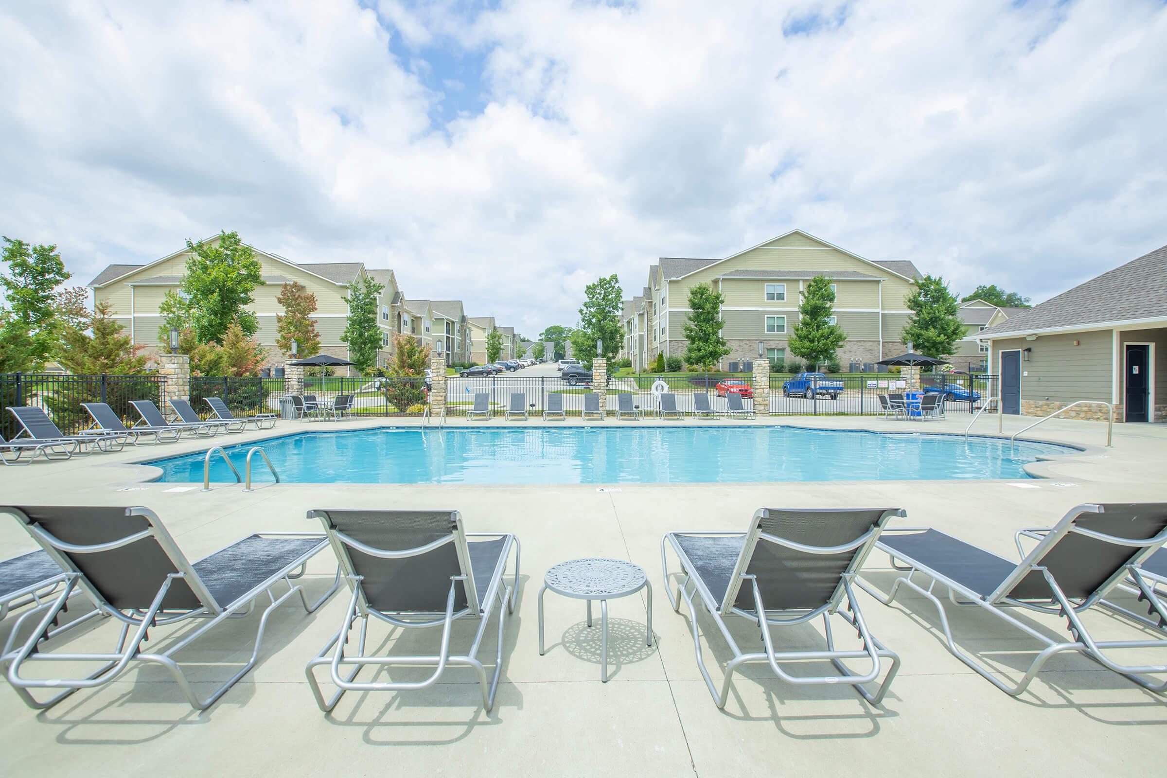 A crystal-clear swimming pool surrounded by lounge chairs under a cloudy sky, with residential buildings and trees in the background.