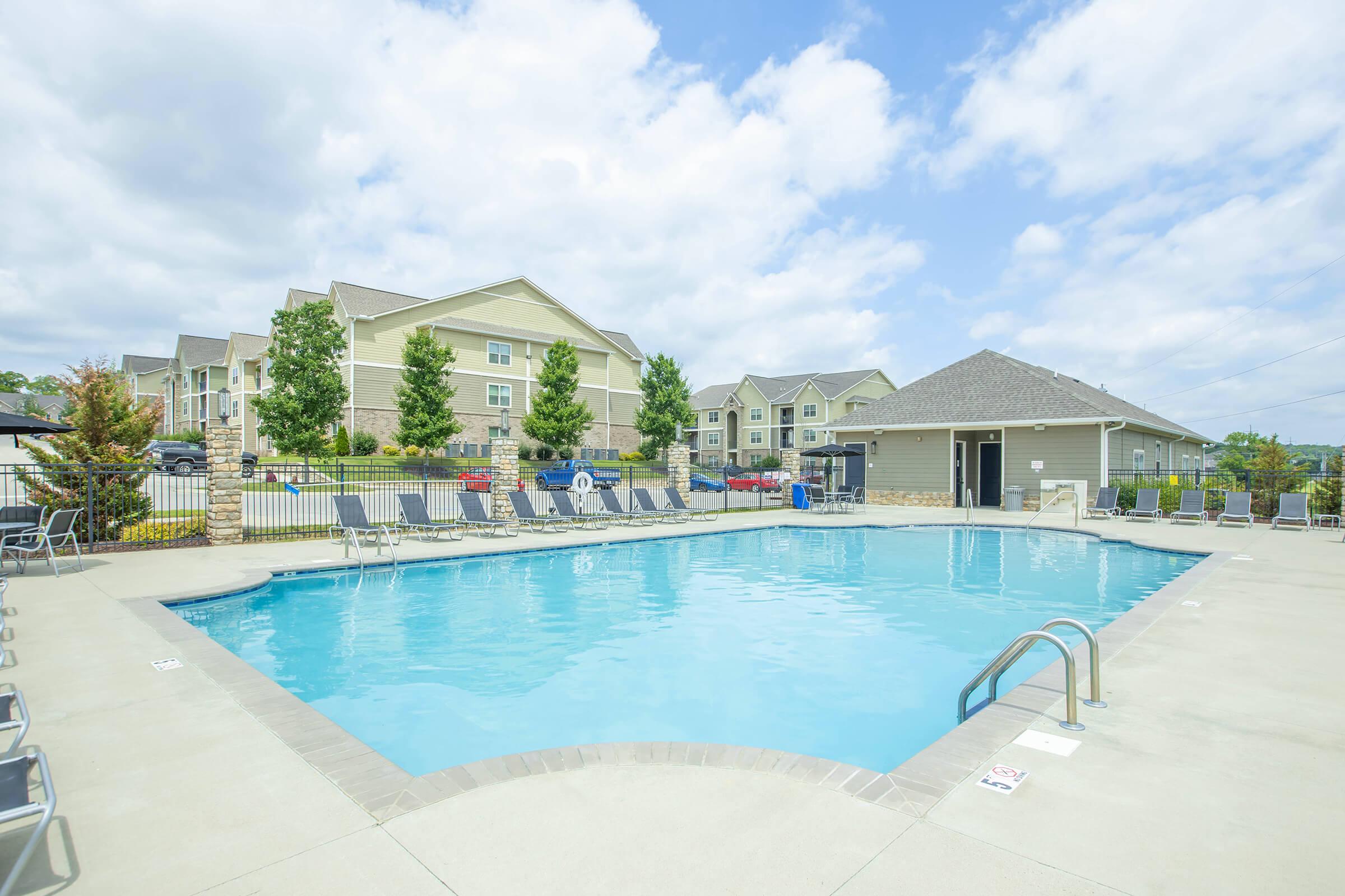 A swimming pool with clear blue water, surrounded by lounge chairs. In the background, there are modern apartment buildings and a cloudy sky.