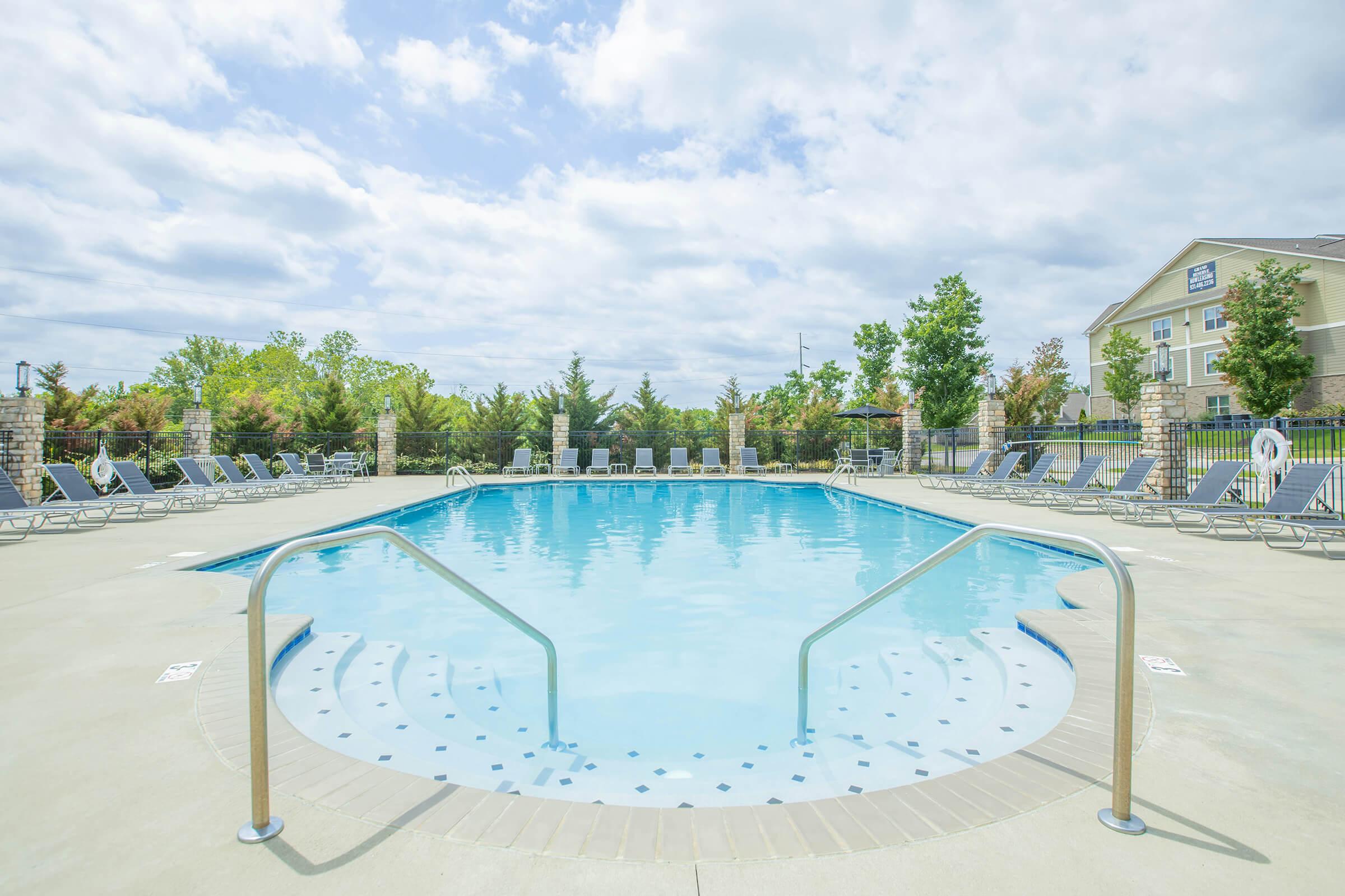 A clear swimming pool surrounded by lounge chairs, with a cloudy blue sky above and trees in the background. The pool features a set of stairs leading into the water.