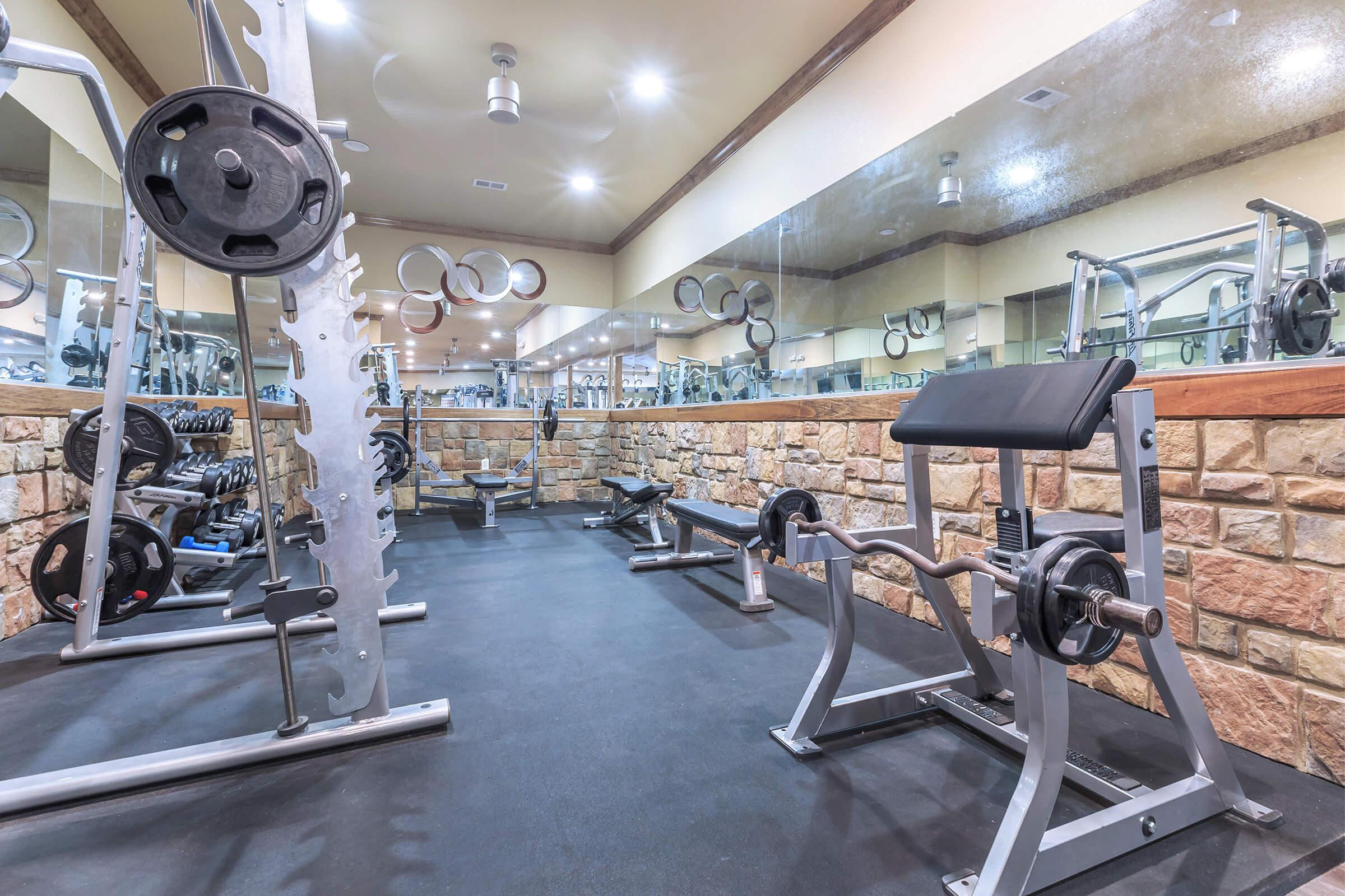 A modern gym interior showcasing weightlifting equipment, including benches and free weights, with stone walls and large mirrors reflecting the workout area.