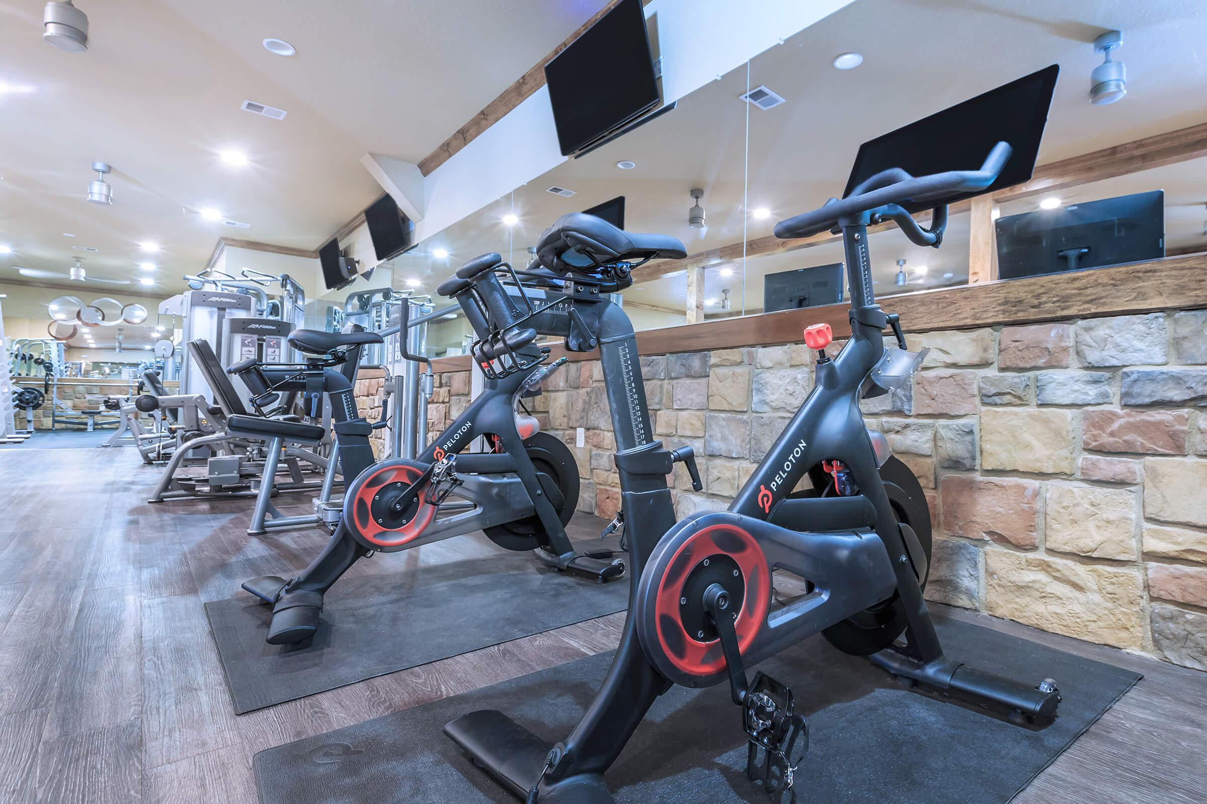 Two stationary exercise bikes positioned in a modern gym with mirrors and stone walls. The gym features additional workout equipment, including weight machines, and is well-lit with several overhead lights.