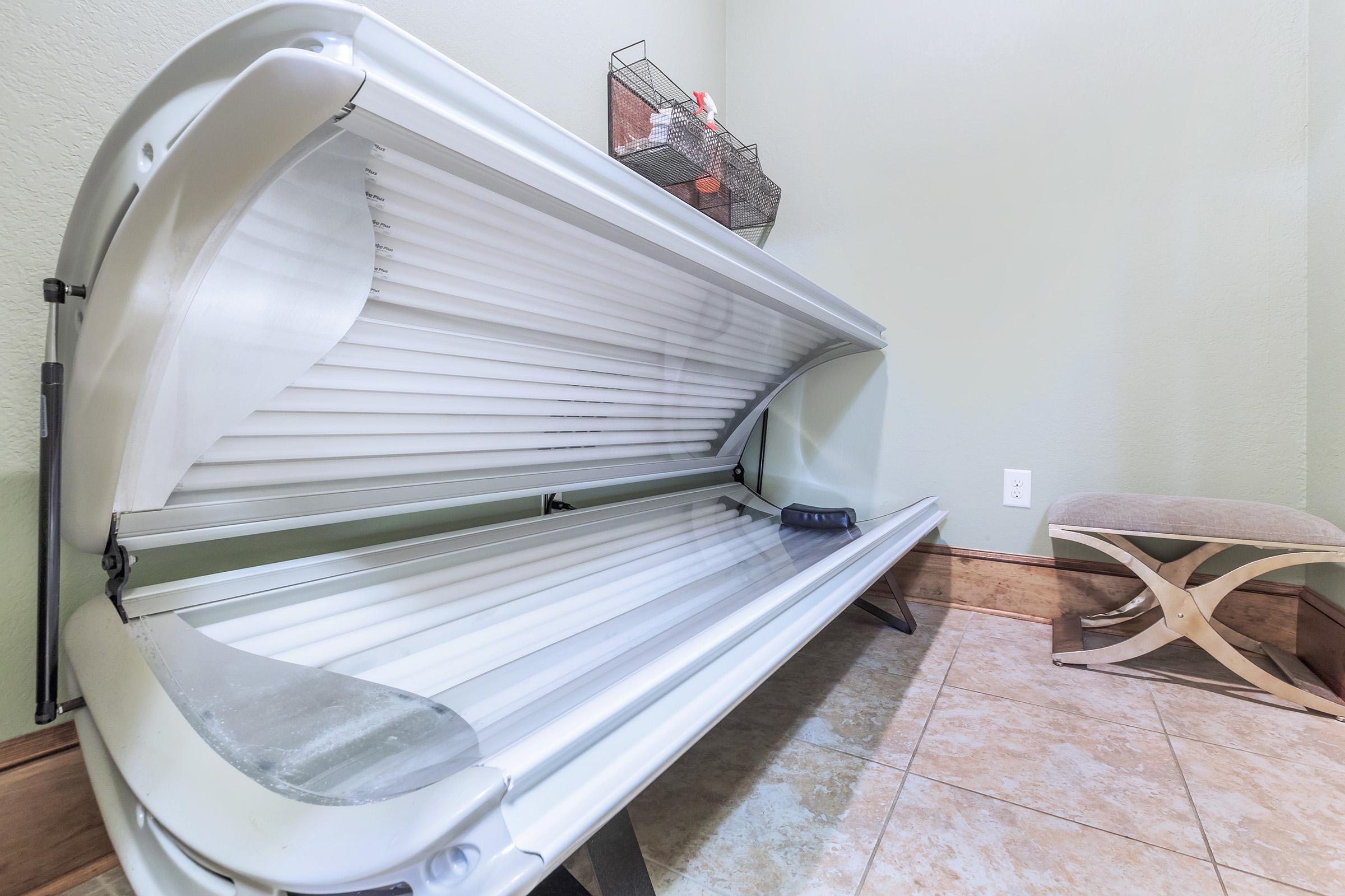 Interior view of a tanning bed in a room with light green walls. A small stool is positioned beside it, and there is a shelf with items in the background. The floor is tiled.