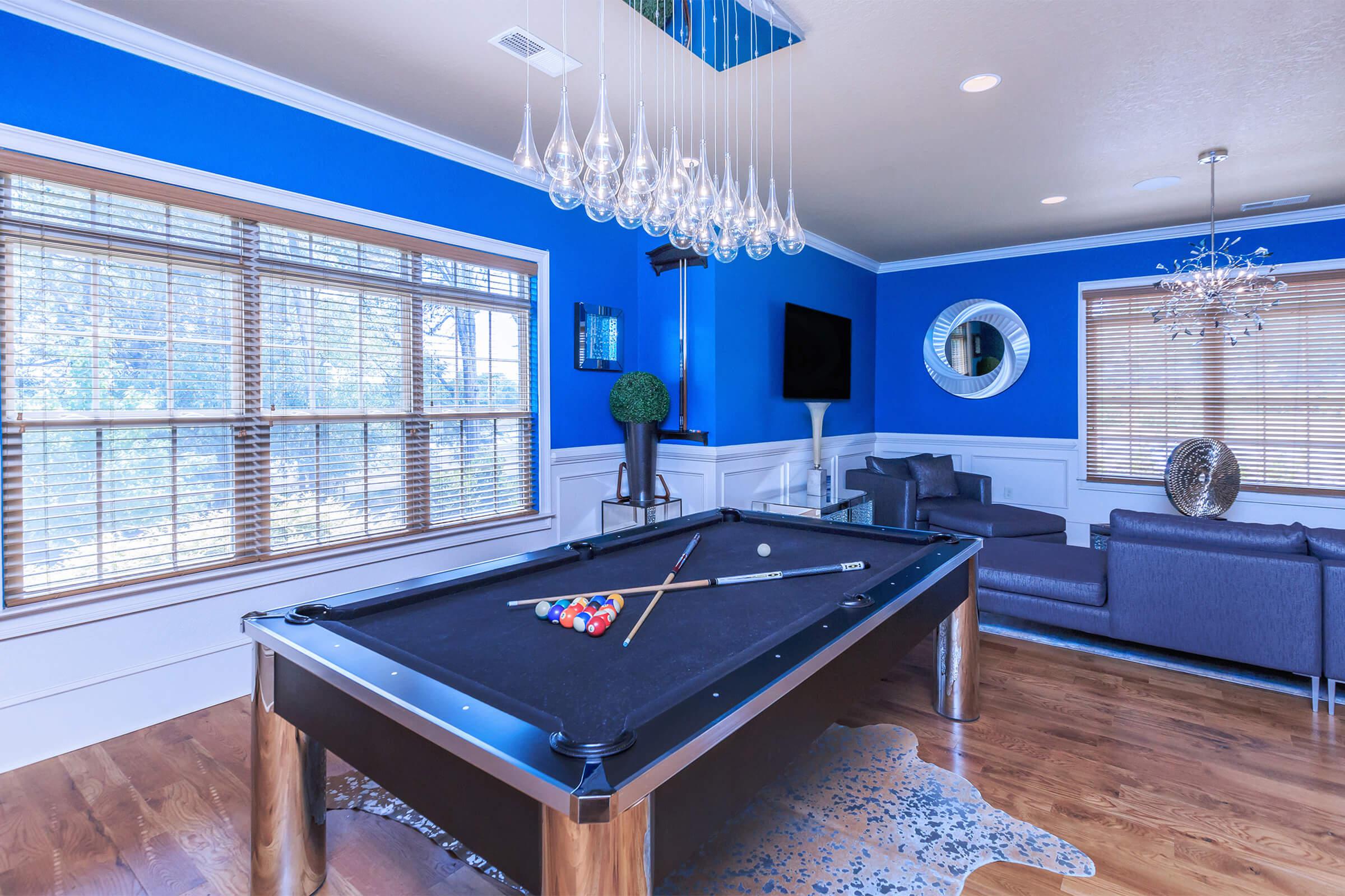 A modern game room featuring a black pool table with colorful balls, a blue accent wall, large windows with wooden blinds, stylish seating, and contemporary lighting fixtures.