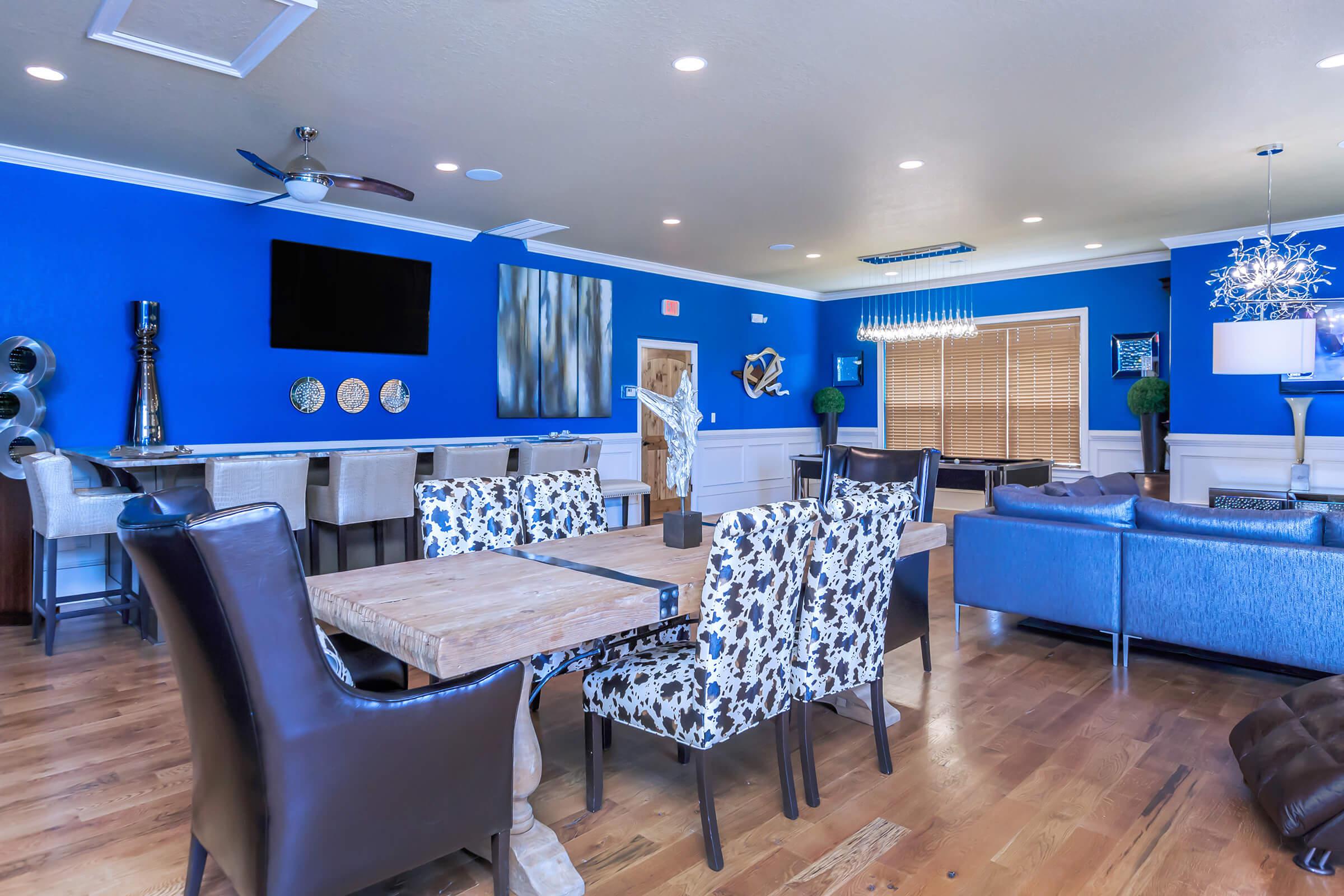 A modern living space with blue walls, featuring a wooden dining table surrounded by patterned chairs, a lounge area with blue sofas, and a bar area in the background. Decorative wall art and a flat-screen TV are visible, along with a light fixture and indoor plants.