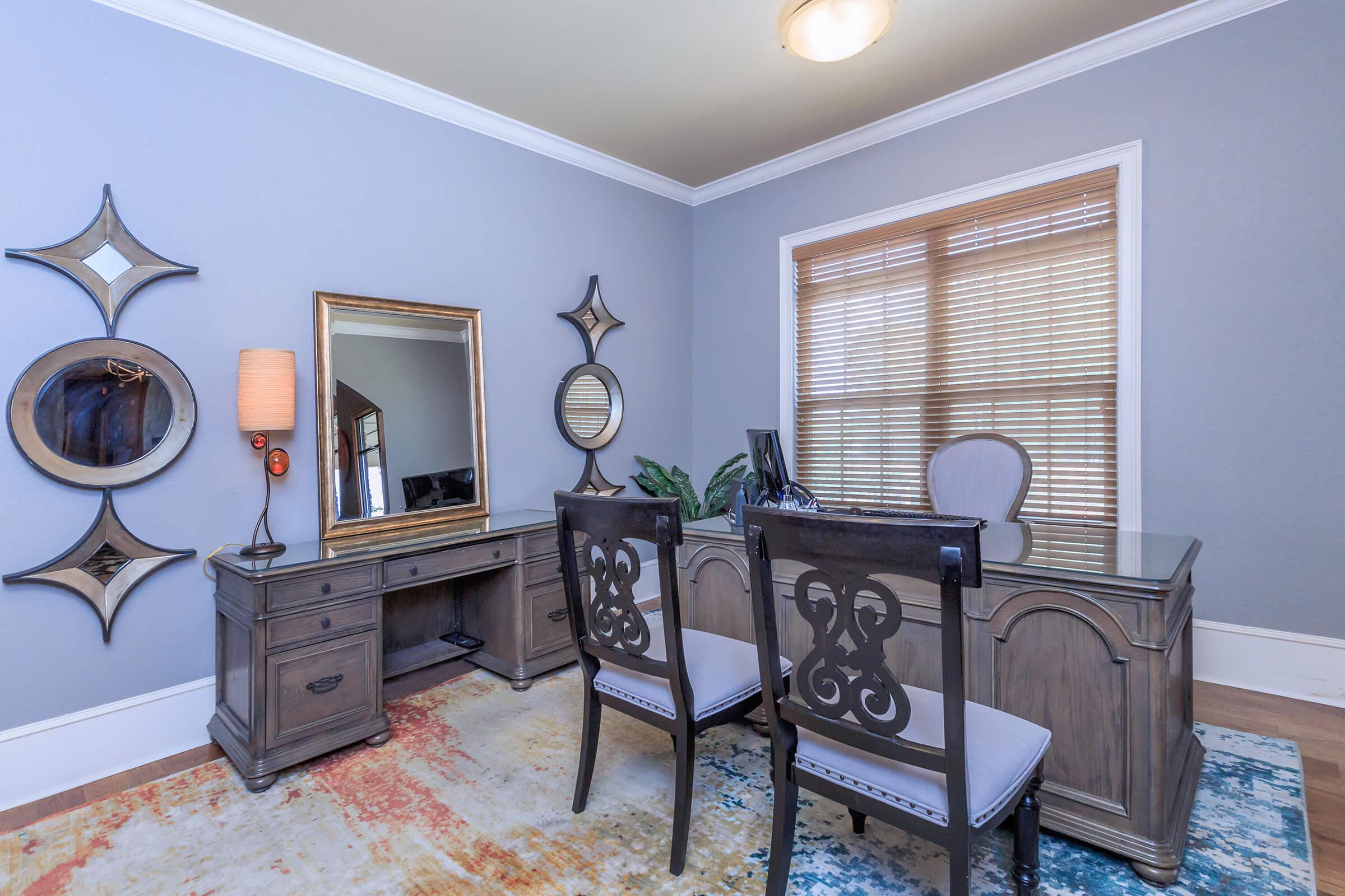 A modern office space featuring a wooden desk and two elegant chairs. A large mirror and decorative wall mirrors are mounted on a light gray wall. There is a potted plant on the desk, and the floor has a colorful area rug. Natural light enters through wooden blinds on the window.