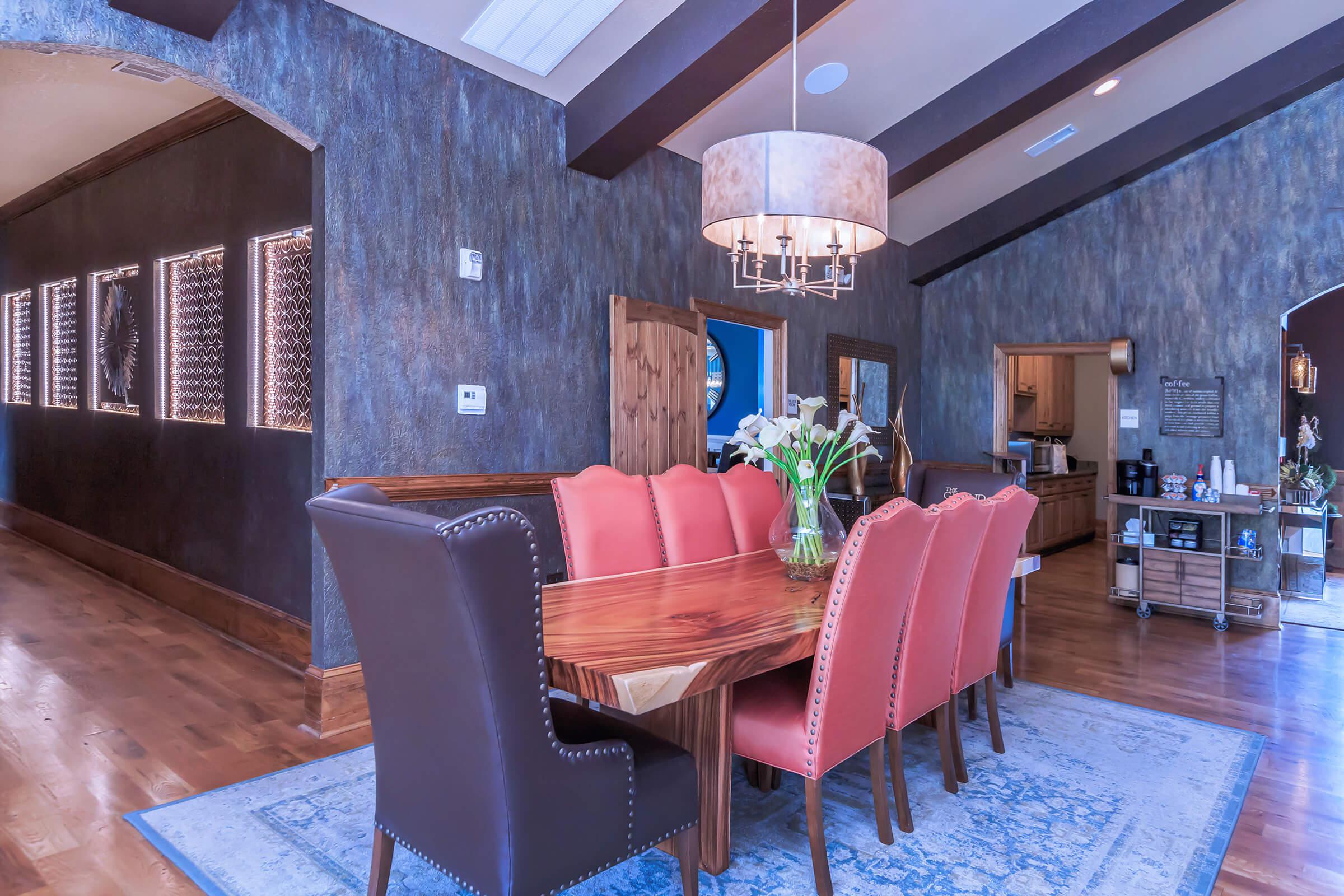 A modern dining room featuring a large wooden table surrounded by stylish chairs, a pendant light fixture above the table, and a textured wall with decorative lighting elements. A vase of flowers sits on the table, and there is a doorway leading to a kitchen area. The flooring is hardwood, and there's a decorative rug beneath the table.