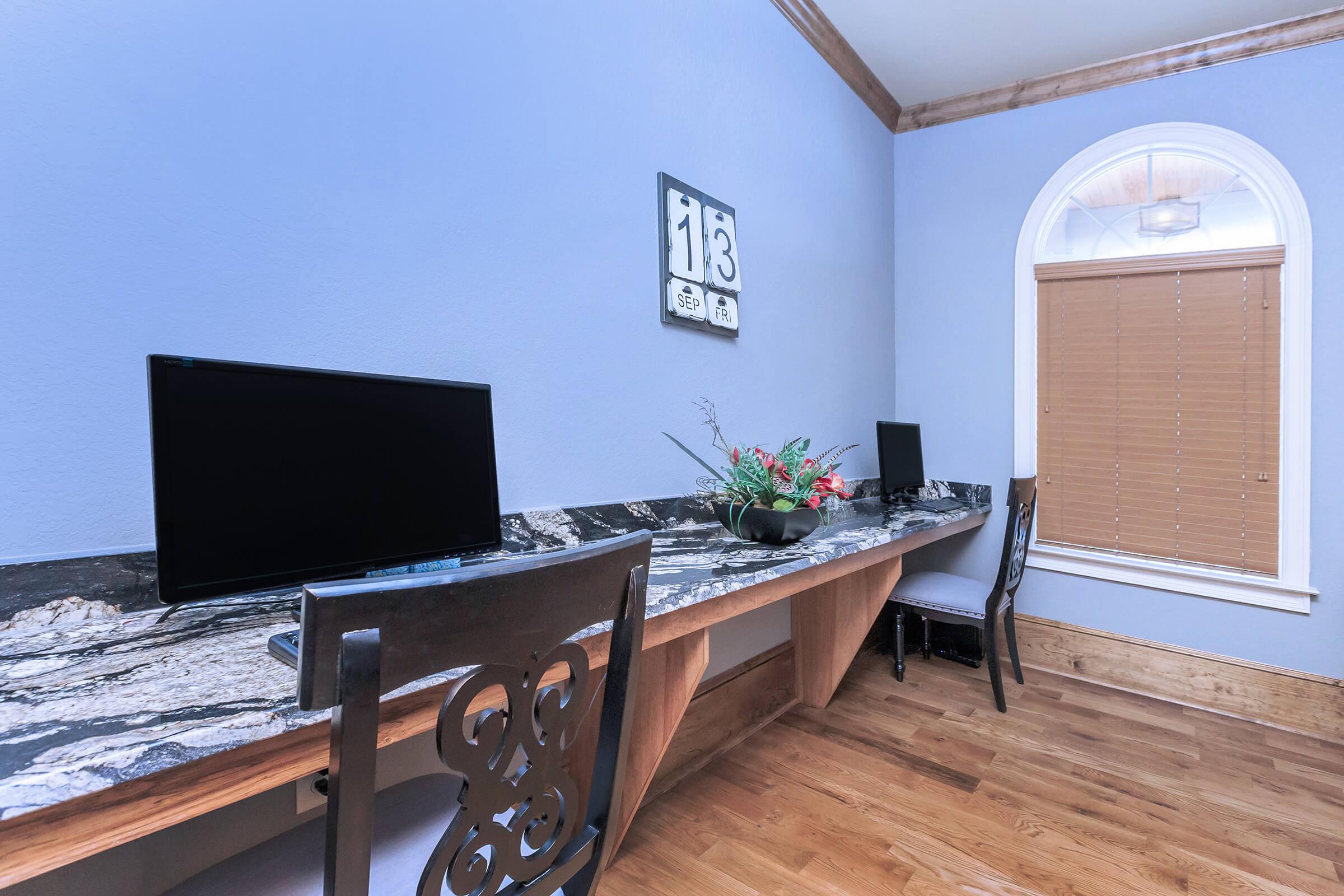 A home office space featuring two black computer monitors on a granite counter, with a decorative plant in the center. The walls are painted light blue, and there is a window with wooden blinds. A wall clock displaying the time is mounted above the counter.