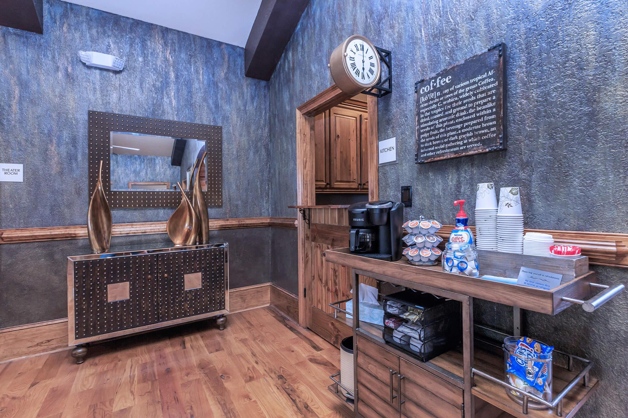 A cozy coffee station in a wooden-themed interior, featuring a coffee maker, cups, a decorative mirror, and a small cart with snacks. A wall clock and a sign about coffee are visible.