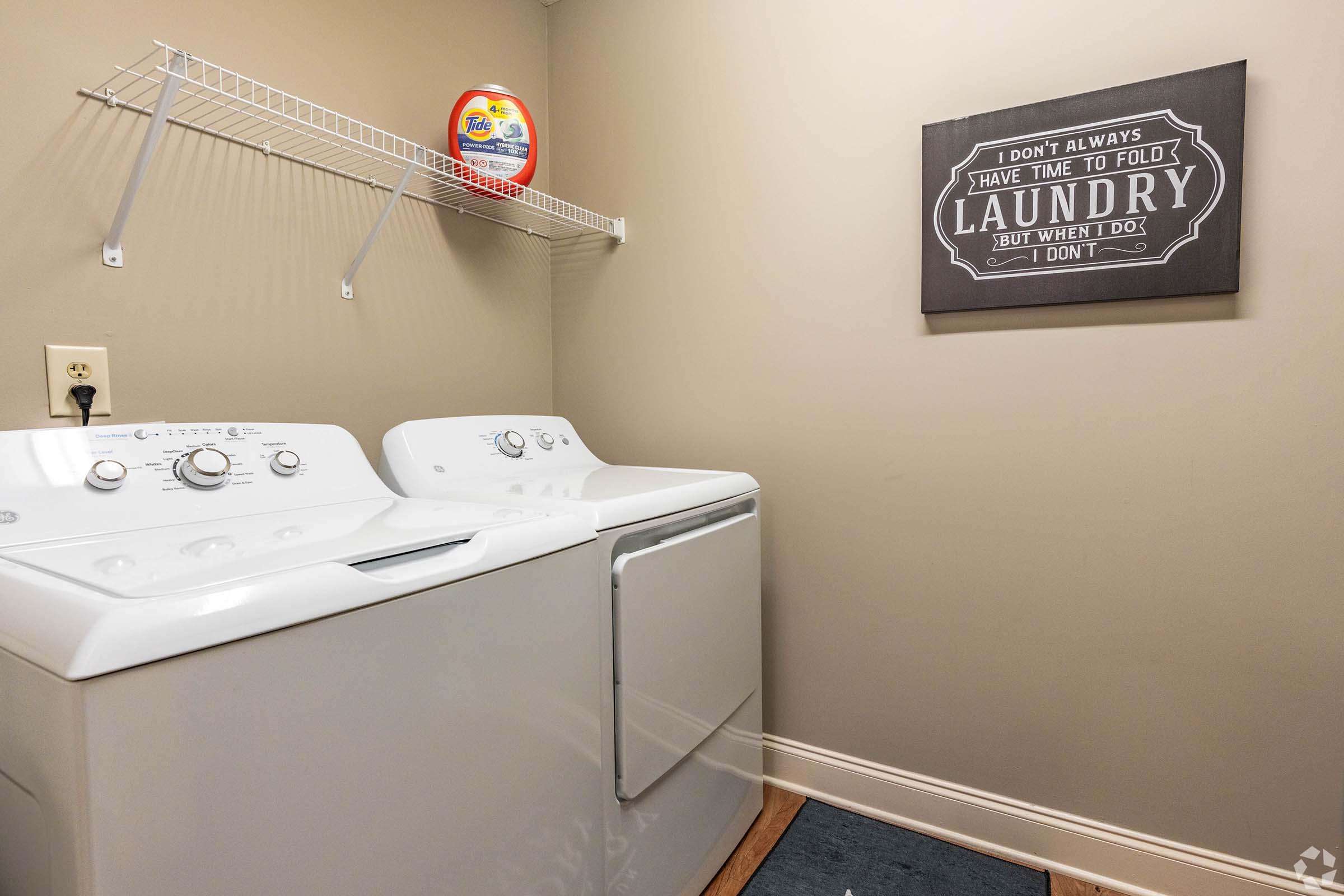 A laundry room featuring a white washing machine and dryer side by side. Above the machines, there's a wall shelf with laundry detergent. A decorative sign on the wall reads, "I DON'T ALWAYS HAVE TIME TO FOLD LAUNDRY BUT WHEN I DO, I DON'T." The walls are painted a neutral color, creating a functional space.
