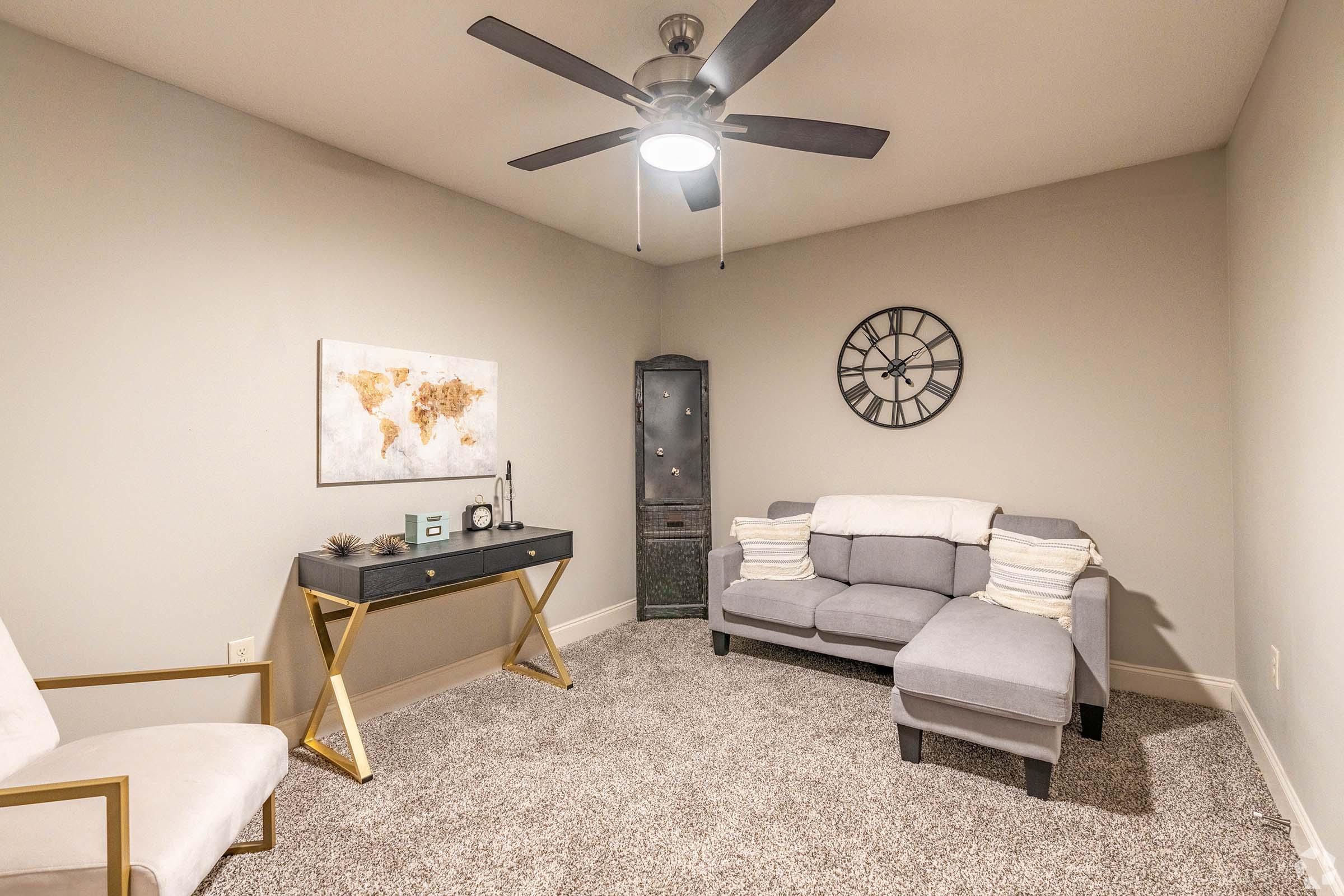 A cozy room featuring a light gray sofa with cushions, a simple white armchair, and a sleek black desk. The walls are painted in a soft color, and a large wall clock and a map art piece add decor. A ceiling fan provides ventilation, and the carpeted floor enhances the room's comfort.