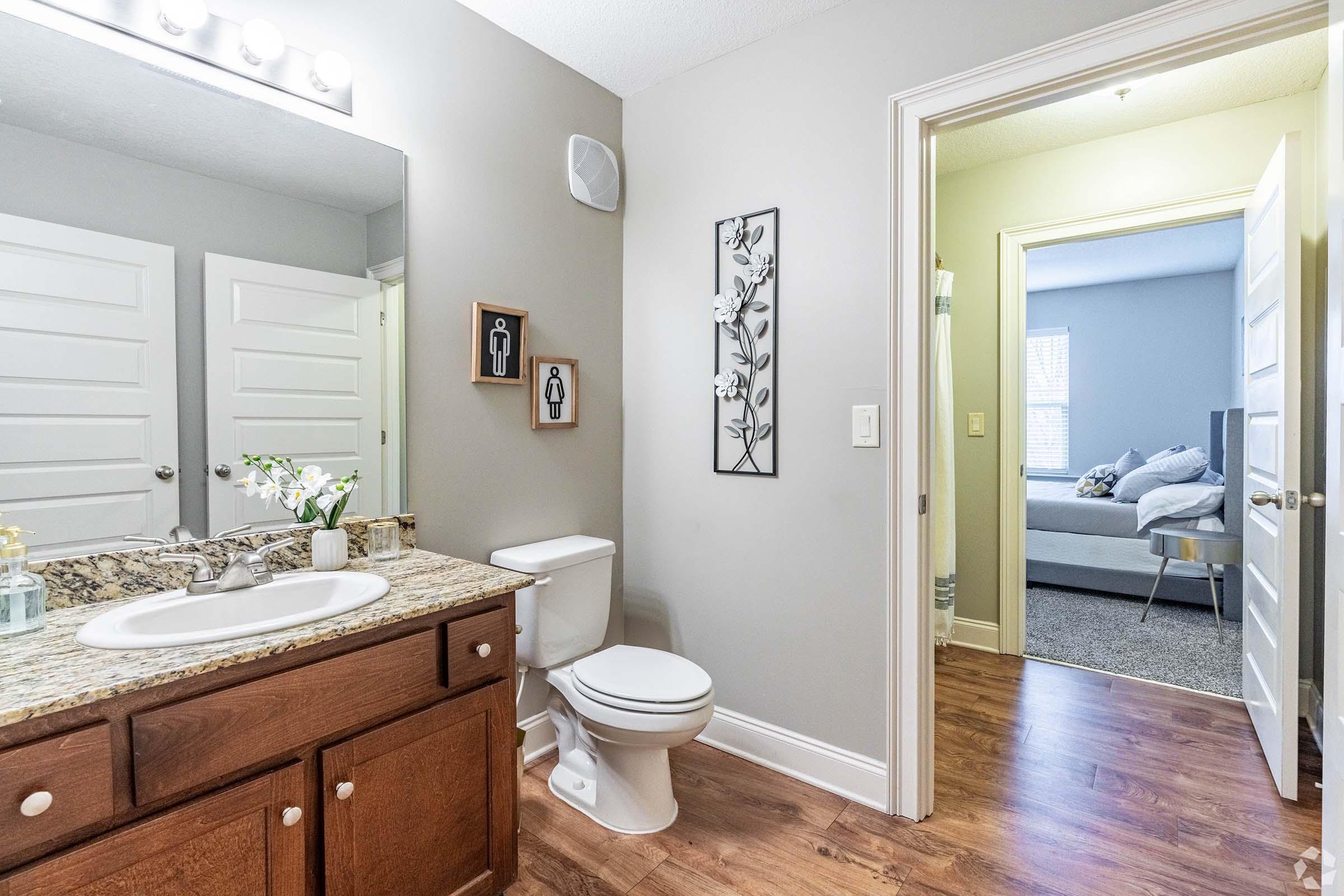 A modern bathroom featuring a granite countertop with a sink and decorative items, a toilet, and stylish wall art. A door leads to a bedroom visible in the background, showcasing a bed and soft textures, while the walls are painted light gray, creating a spacious and inviting atmosphere.