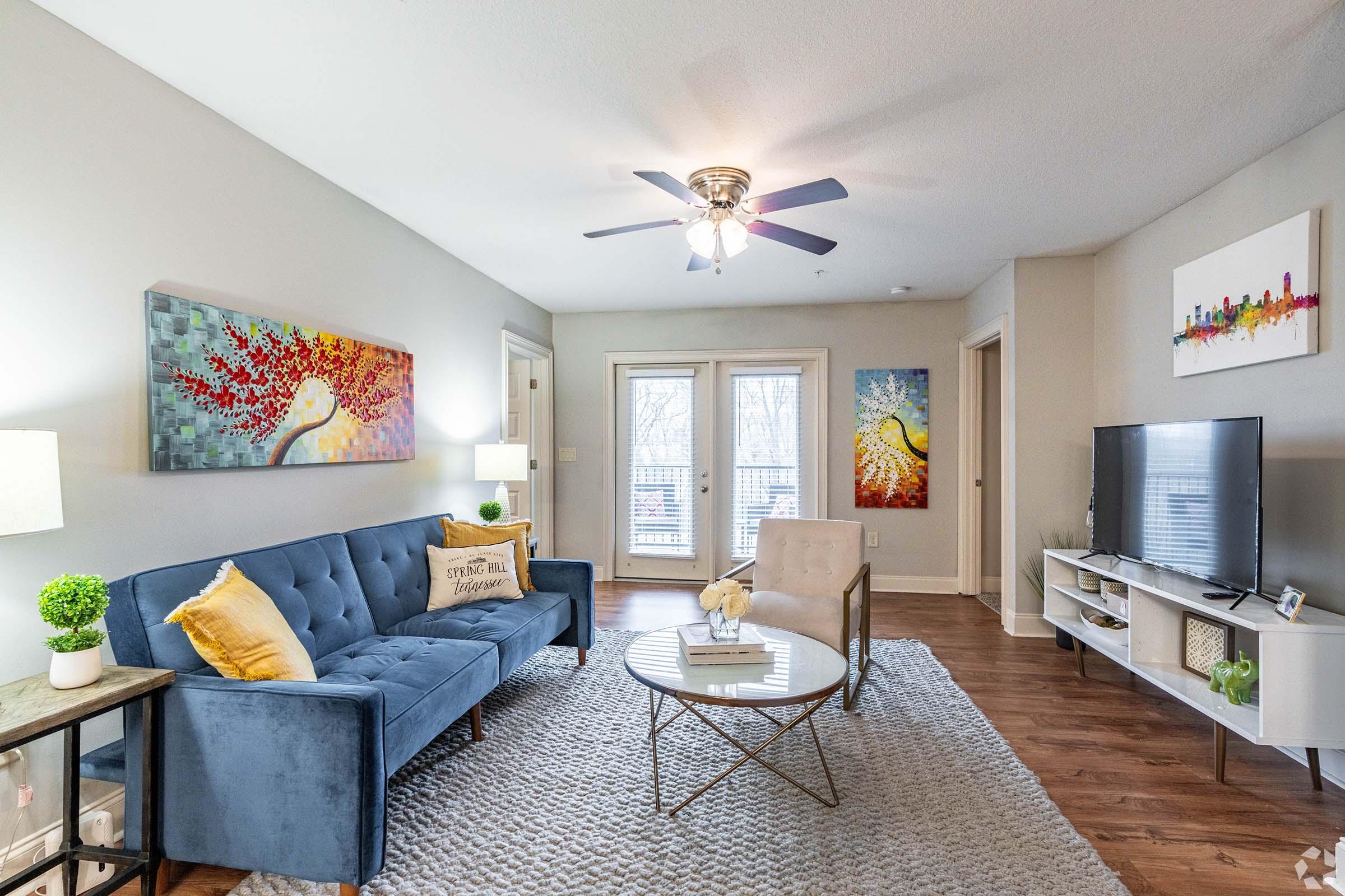 A modern living room featuring a blue sofa with decorative pillows, a round coffee table, and a flat-screen TV on a white stand. The walls are adorned with colorful artwork, and there are large windows with light-colored curtains. A soft rug covers the wooden floor, and potted plants add a touch of greenery.