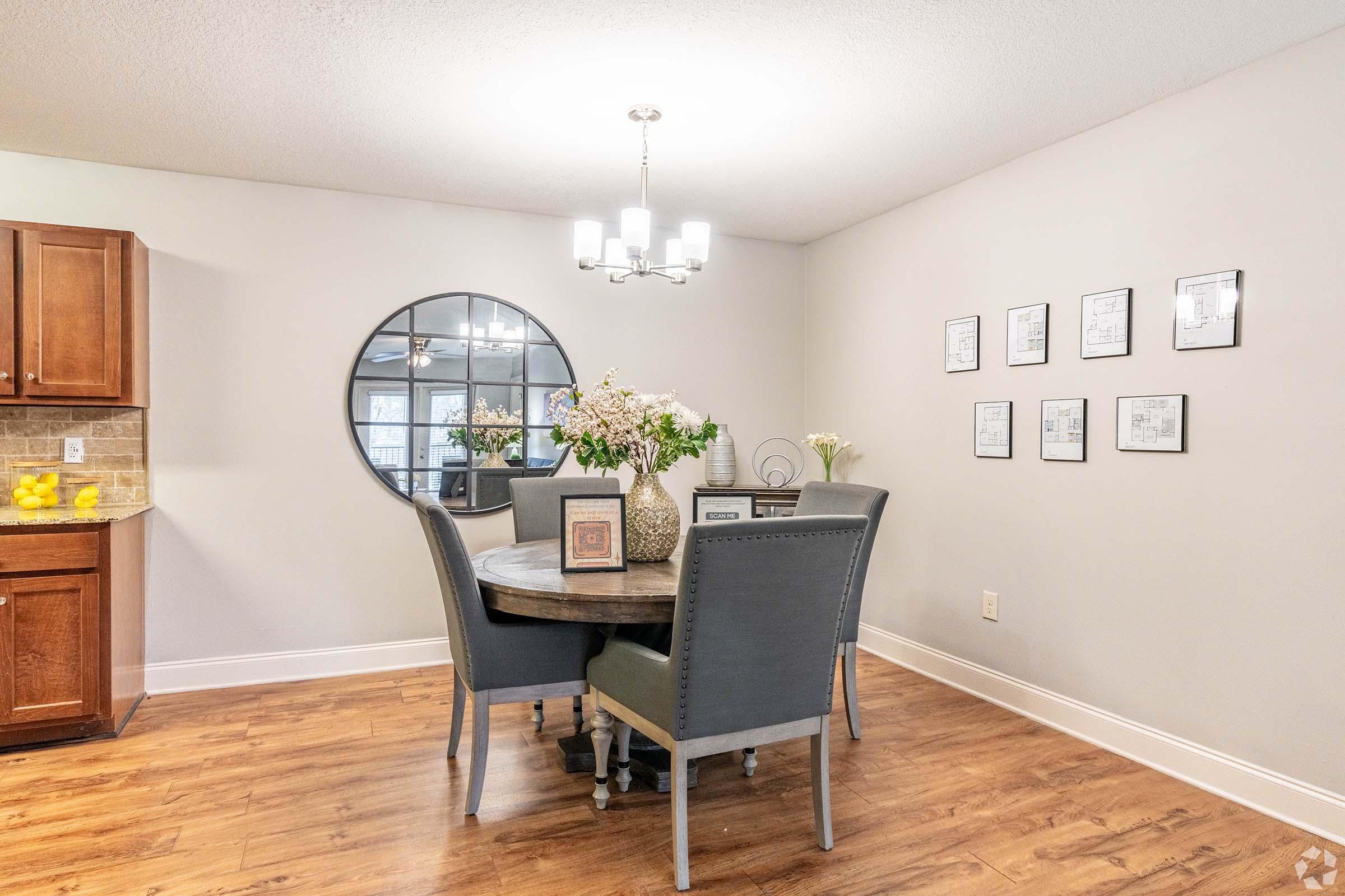 A modern dining area featuring a round wooden table surrounded by four gray chairs. A circular mirror hangs on the wall, reflecting light from a stylish chandelier above. The walls are adorned with framed artwork, and a kitchen area with wooden cabinets is visible in the background.