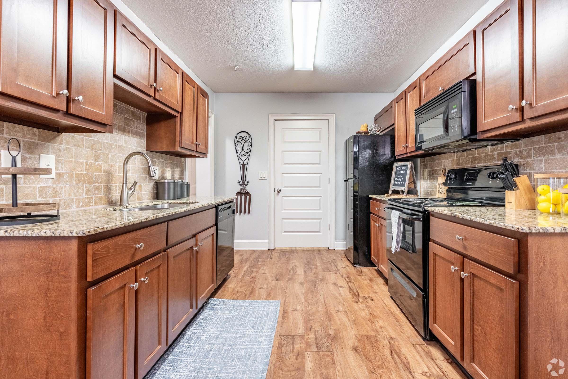 Modern kitchen featuring dark wood cabinets, granite countertops, and stainless steel appliances. The space is well-lit with a ceiling fixture, showcasing a clean and organized layout. A door leads to another room, with a decorative wall hanging visible.