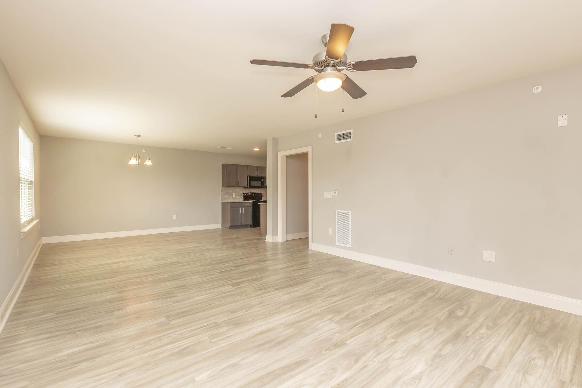 Spacious living area with light gray walls and laminate flooring. A ceiling fan with a light fixture is centered in the room. In the background, a kitchen with dark cabinetry and stainless steel appliances is visible. Large windows allow natural light to fill the space.