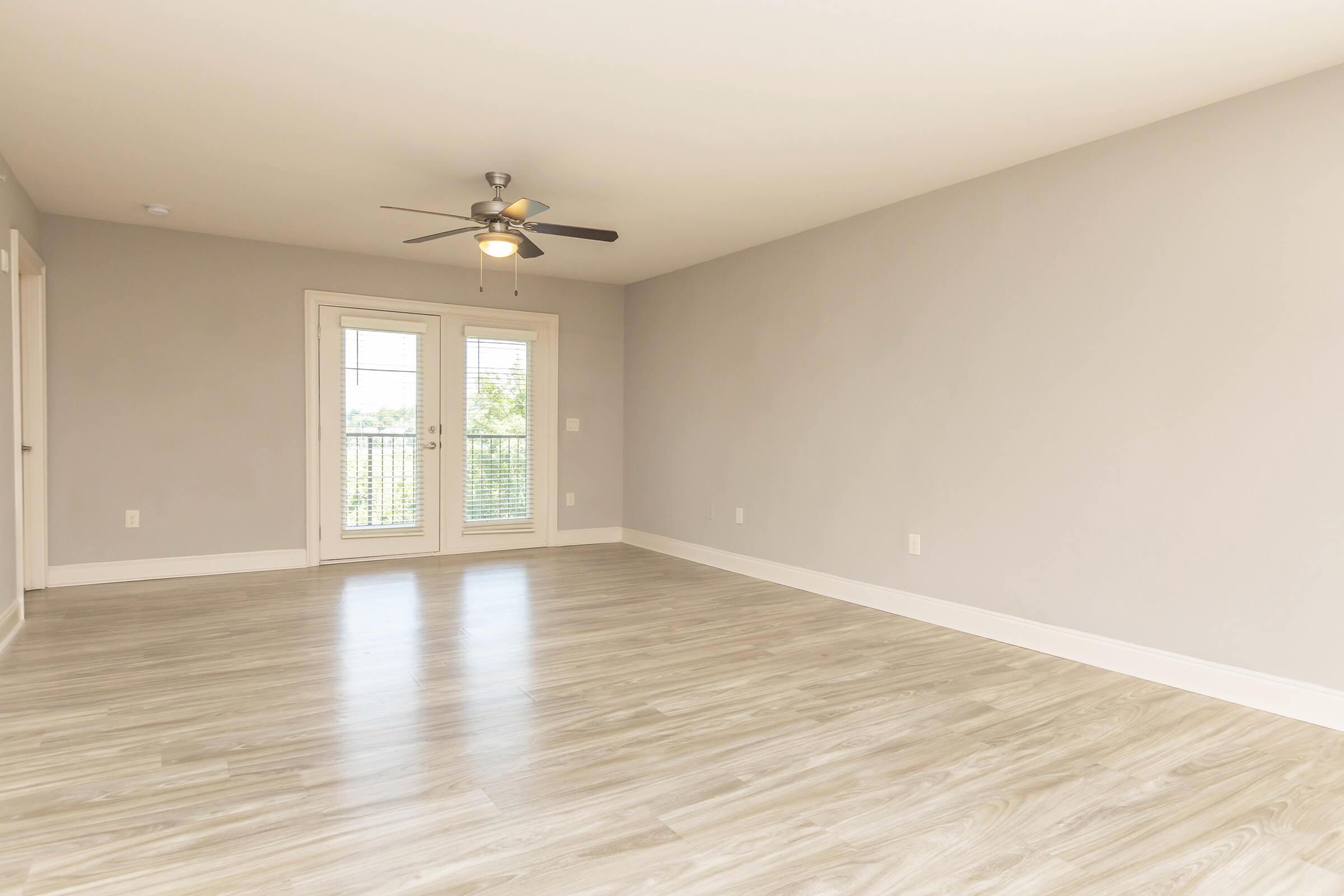 A spacious room with light-colored walls and a smooth wooden floor. There is a ceiling fan visible, and large windows with blinds that allow natural light. The room appears empty, offering a clean and open space suitable for various furnishings or purposes.