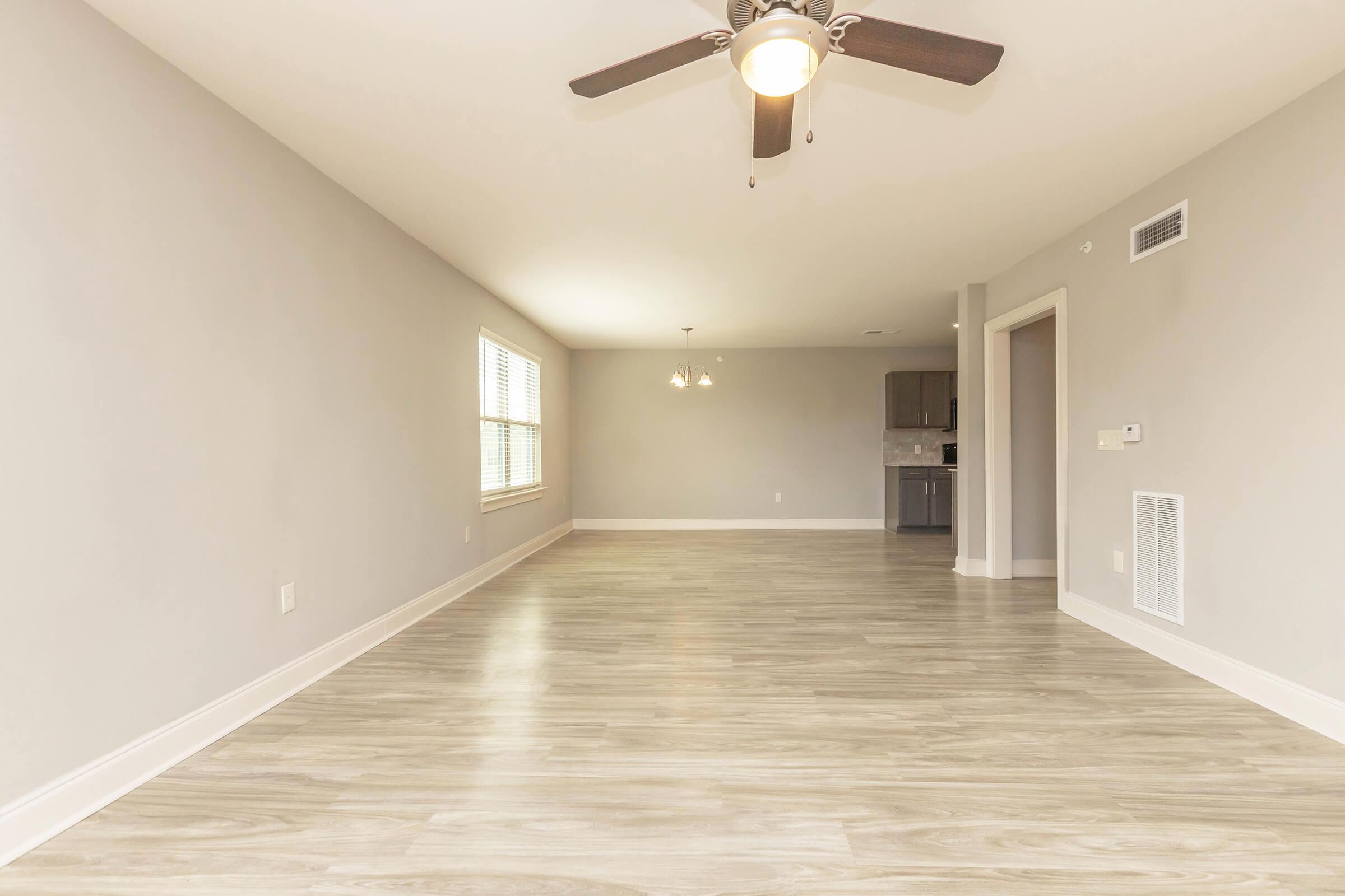 A spacious, well-lit living room featuring light gray walls and a light wood floor. There is a ceiling fan and large windows providing natural light. In the background, a dining area can be seen leading to a kitchen, creating an inviting and open atmosphere.