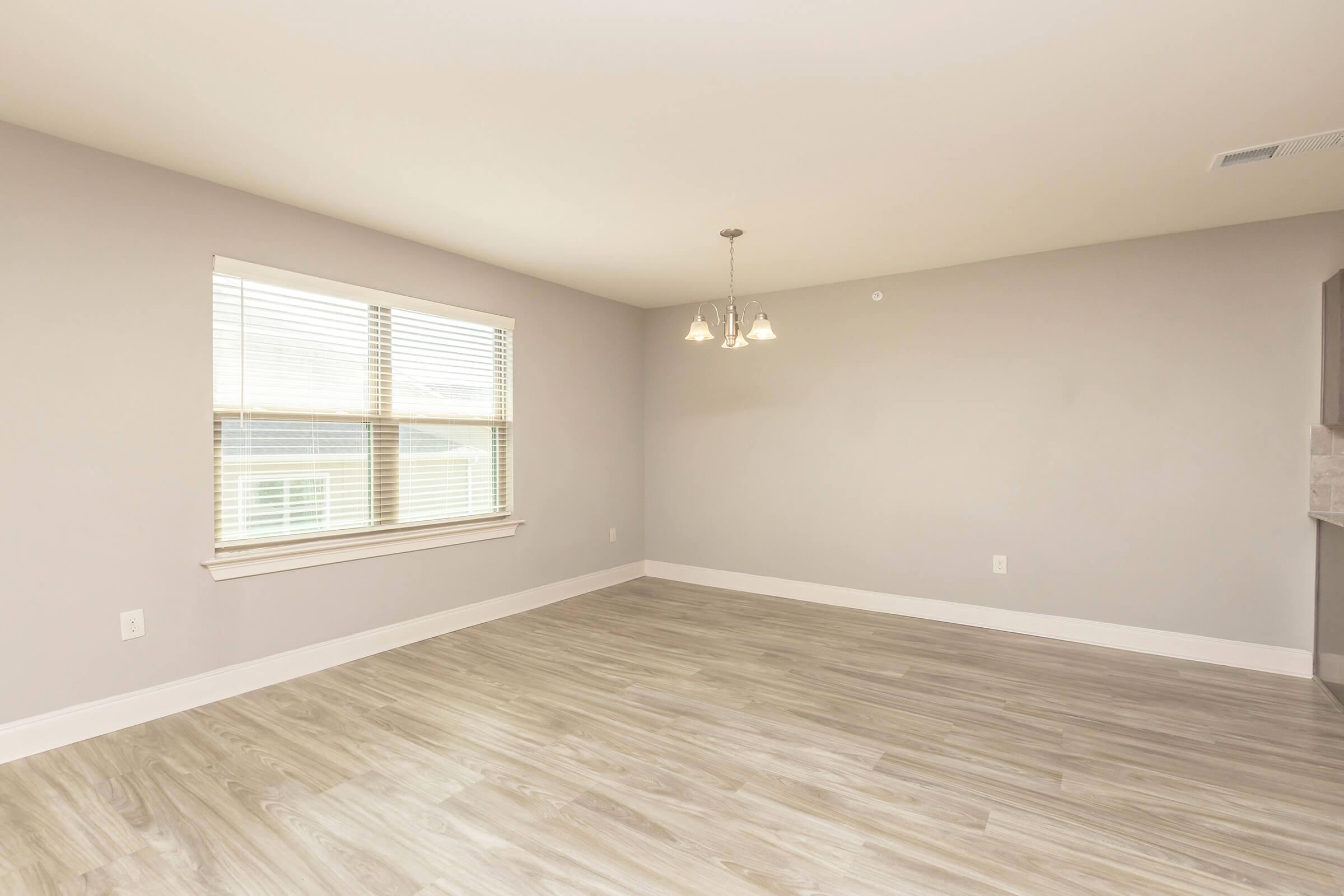 A spacious, empty room with light gray walls and wood-look flooring. There is a window with horizontal blinds allowing natural light, and three pendant lights hanging from the ceiling. The room has a clean, modern aesthetic and provides ample space for furniture arrangement.