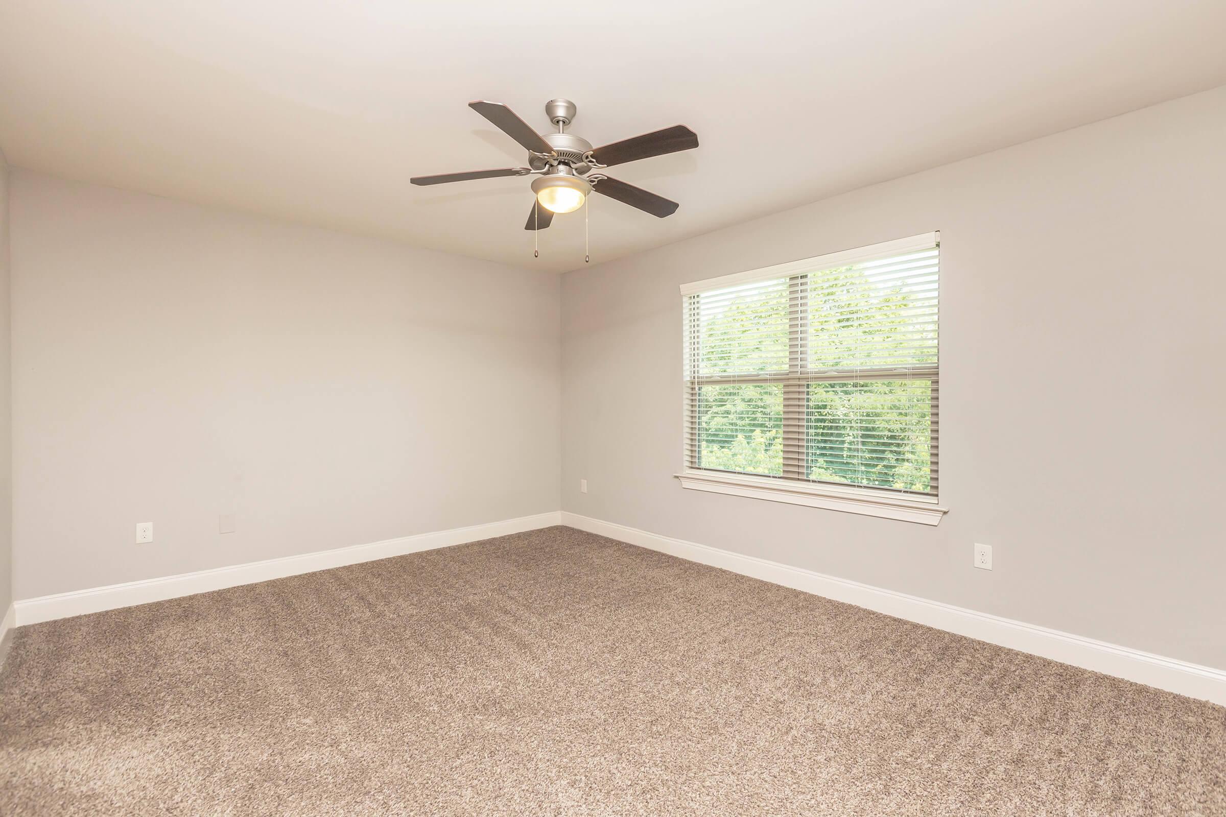 A spacious, empty room with light gray walls and soft carpet flooring. It features a ceiling fan with five blades and a window with blinds, allowing natural light to fill the space. The overall atmosphere is bright and clean, ideal for personal customization.