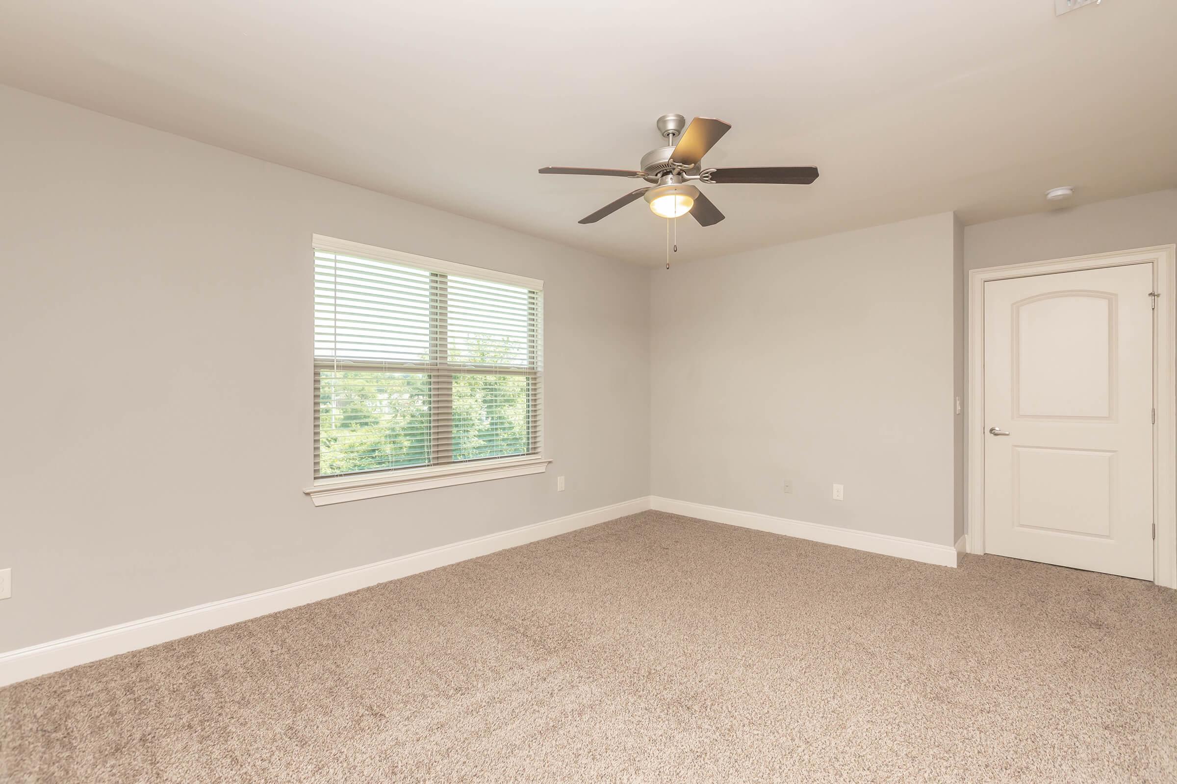 A spacious, empty room with light gray walls and carpeted flooring. It features a ceiling fan and a window with closed blinds, allowing natural light to enter. A door is visible on the right side, leading to another area. The space is clean and well-lit, suitable for various uses like a bedroom or an office.