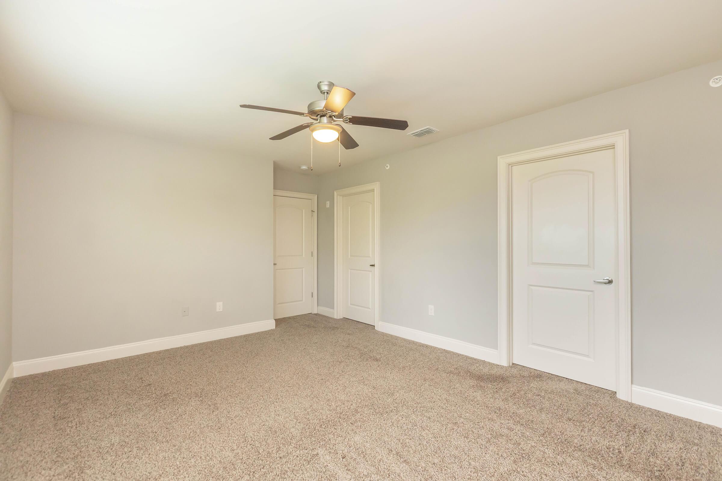 A light, airy bedroom featuring beige carpet and light gray walls. A ceiling fan with a light fixture is visible. There are two closed doors, one leading to a closet and the other to a hallway. The room is empty, offering a spacious feel and plenty of natural light.