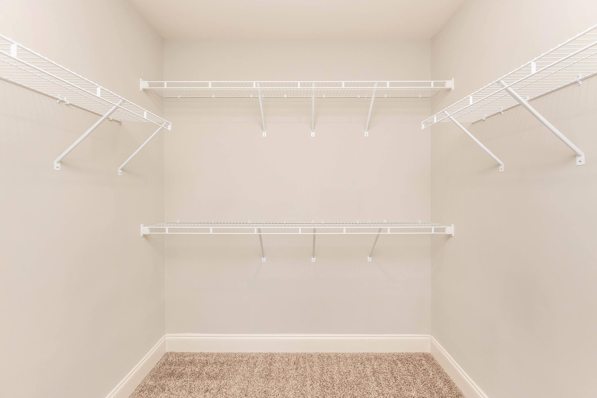 Empty closet with beige walls and a carpeted floor. The closet features metal wire shelving on both sides, providing several levels for storage. The shelves are white and extend from the left side to the right, creating an open and spacious feel.