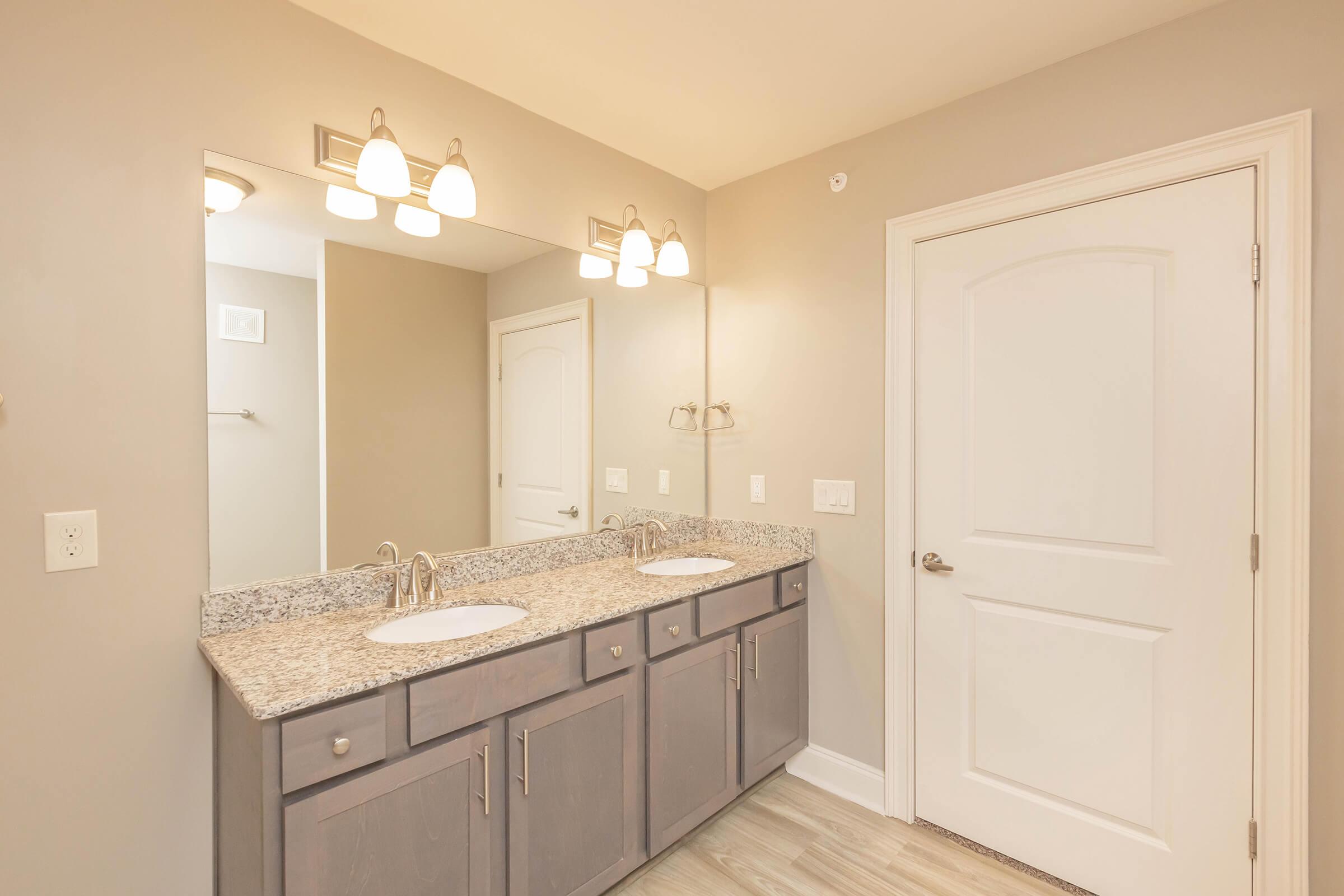 A modern bathroom featuring a double vanity with granite countertops, two sinks, and a large mirror above. The light fixtures are stylishly arranged above the mirror. The walls are painted in a soft neutral tone, and there is a door visible on the right leading to another room. The floor has light-colored wood-like tiles.