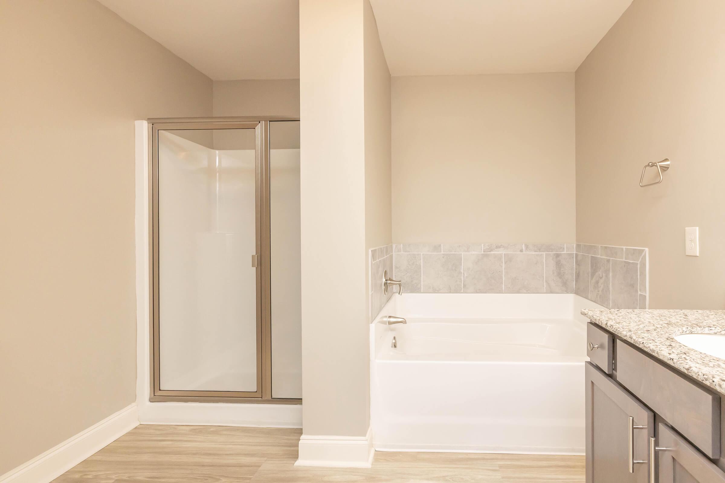 A modern bathroom featuring a glass-enclosed shower on the left, a white bathtub with a tiled surround on the right, and a granite countertop with a sink. The walls are painted in a neutral color, and the flooring is light, giving the space a clean and contemporary look.