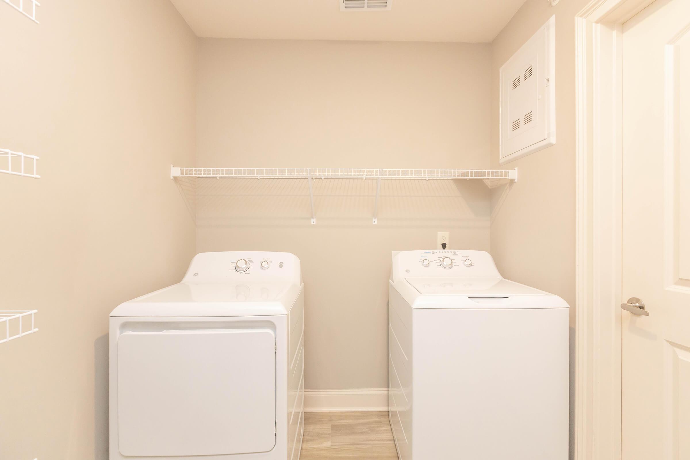 A bright laundry room featuring two white washing machines side by side. The walls are painted a light neutral color, and there is a white wire shelf above the machines for storage. A clean, modern design with tile flooring and a door on the right side.