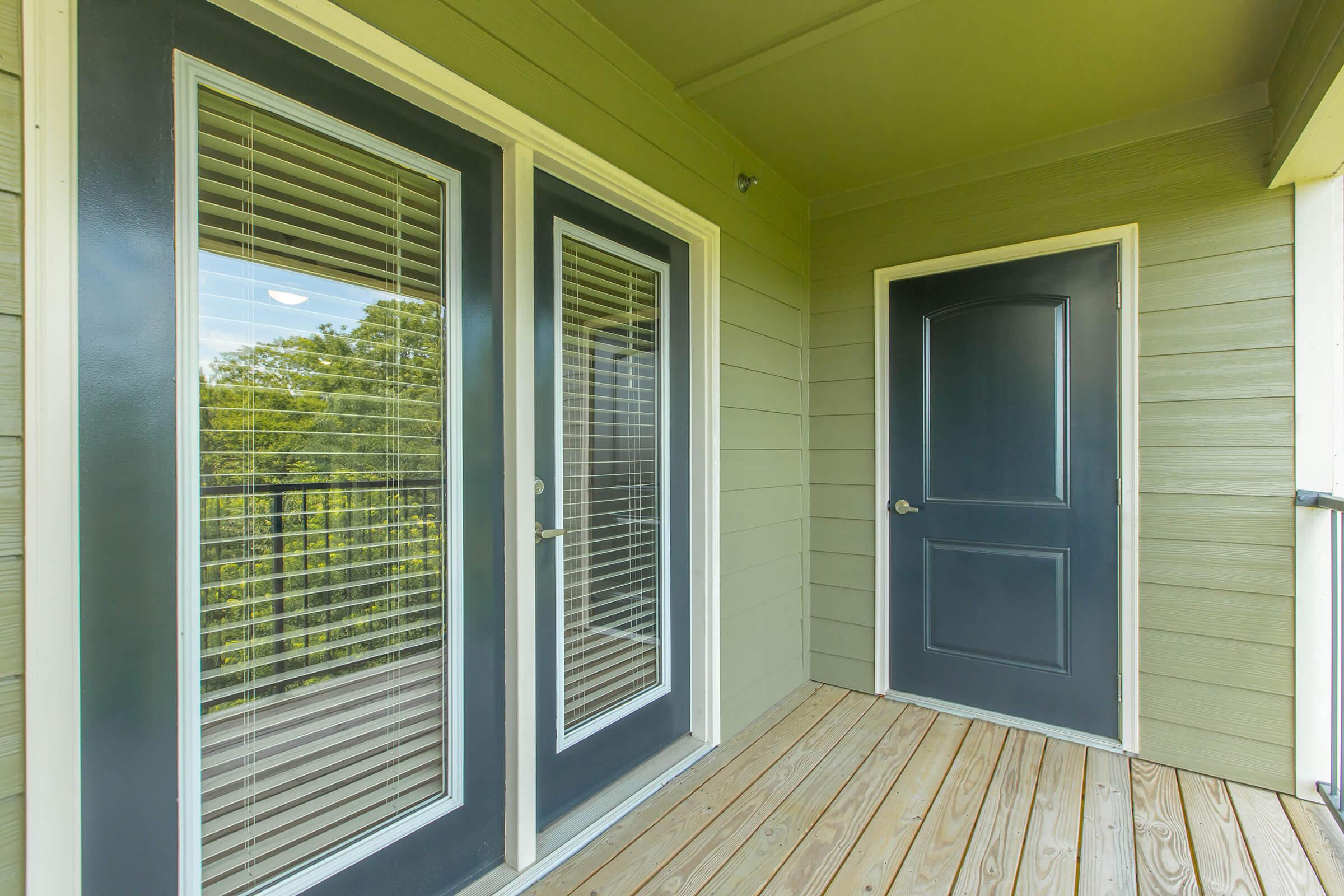 A covered entrance with two glass-paneled doors and a solid door, all framed in a neutral-colored wall. The floor is made of wooden planks, and there are green trees visible through the glass doors, creating a serene outdoor view.